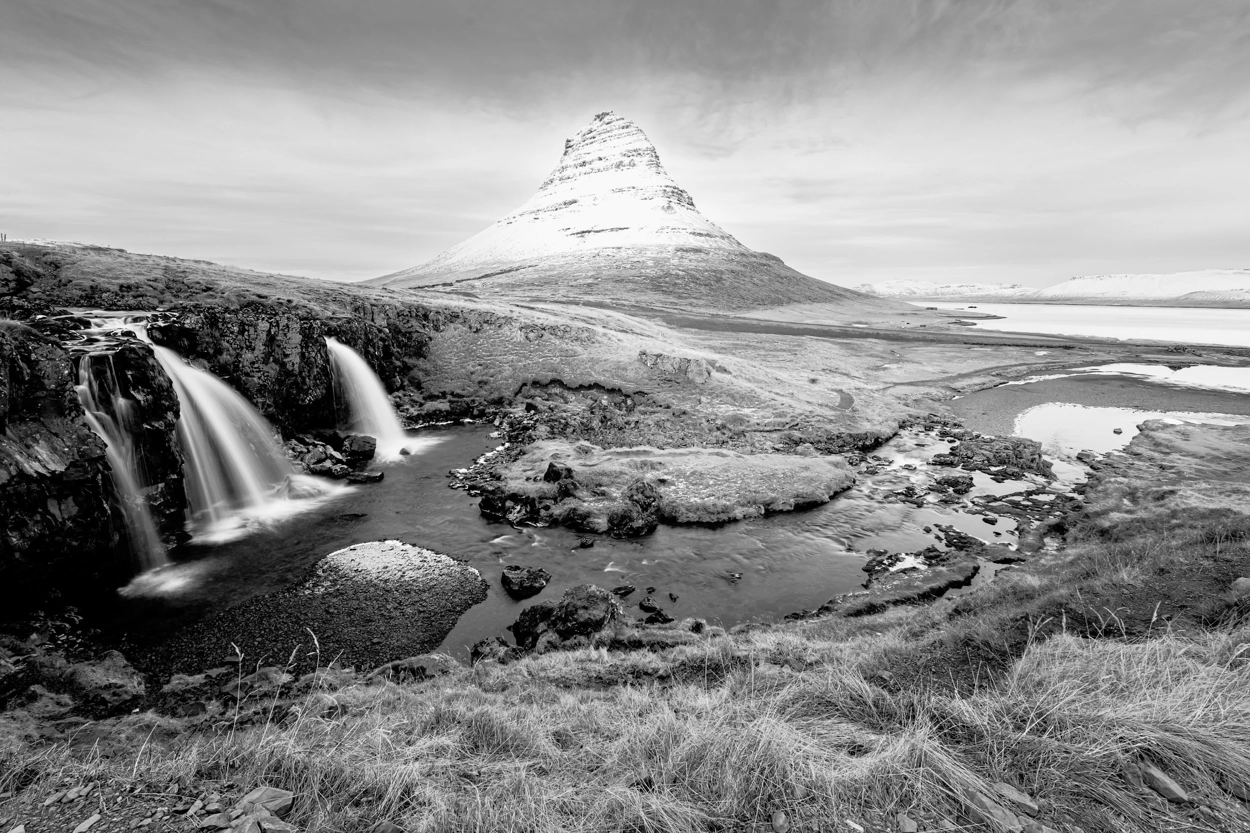Black and white landscape featuring Kirkjufell mountain in Iceland, with waterfalls and streams in the foreground, grassy terrain, and snow-capped peaks in the distance.