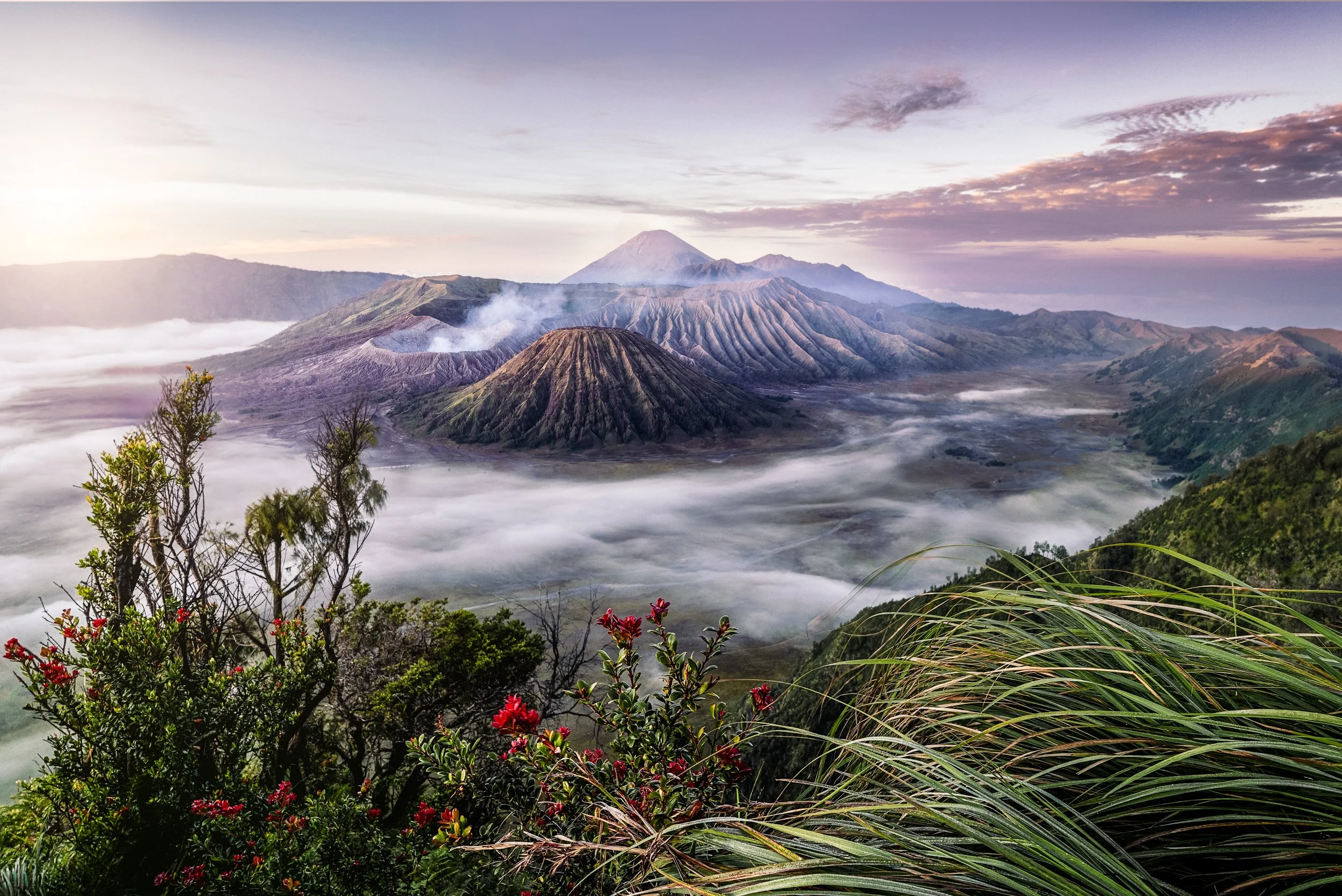 A scenic view of volcanic mountains with ash and smoke, surrounded by lush greenery and clouds, during sunrise or sunset.