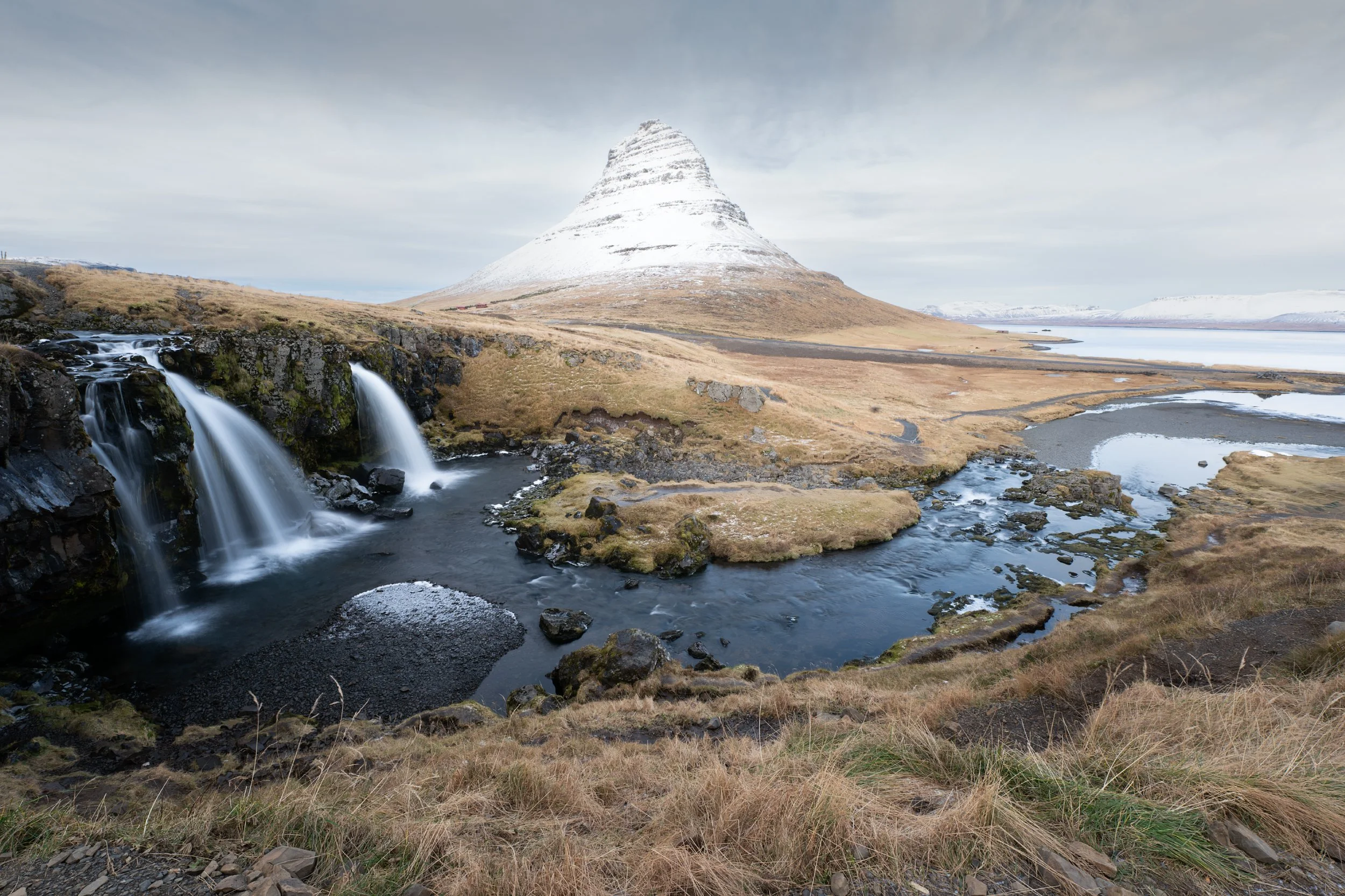 Scenic view of Kirkjufell mountain with a waterfall and river in the foreground, snow on the mountain, and a cloudy sky.