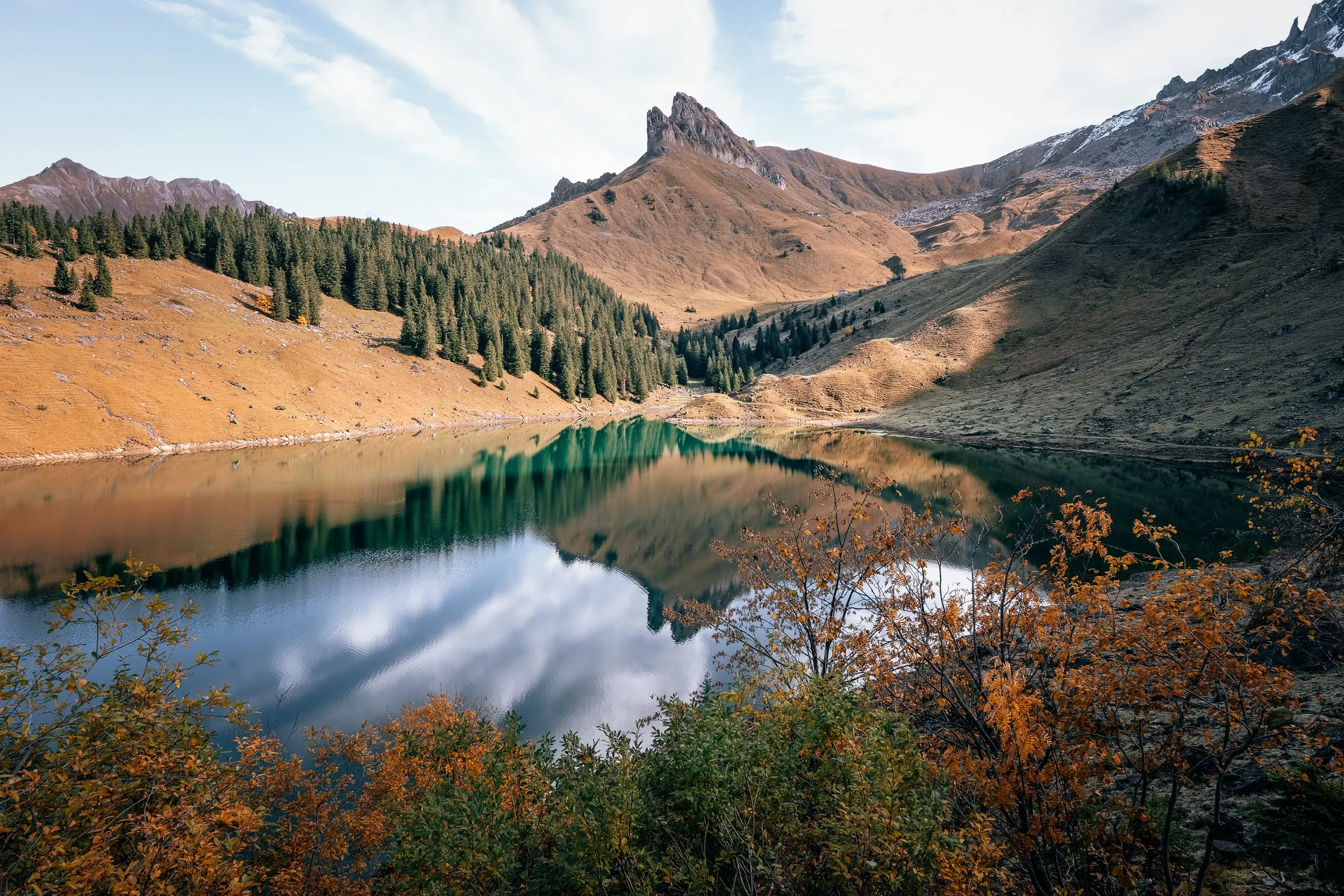 A mountain landscape with a lake in the foreground, reflecting the surrounding mountains and sky, with autumn foliage in the front.