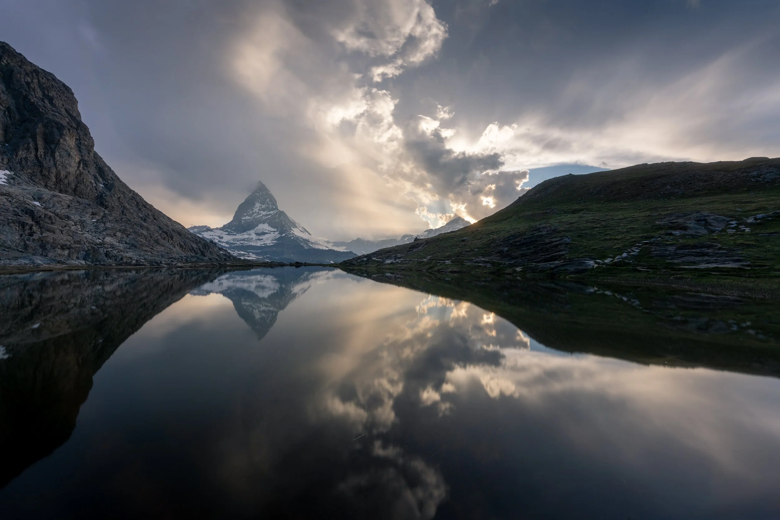 Mountain landscape with Matterhorn peak, water reflecting the sky and mountains, and cloudy sky at dusk.