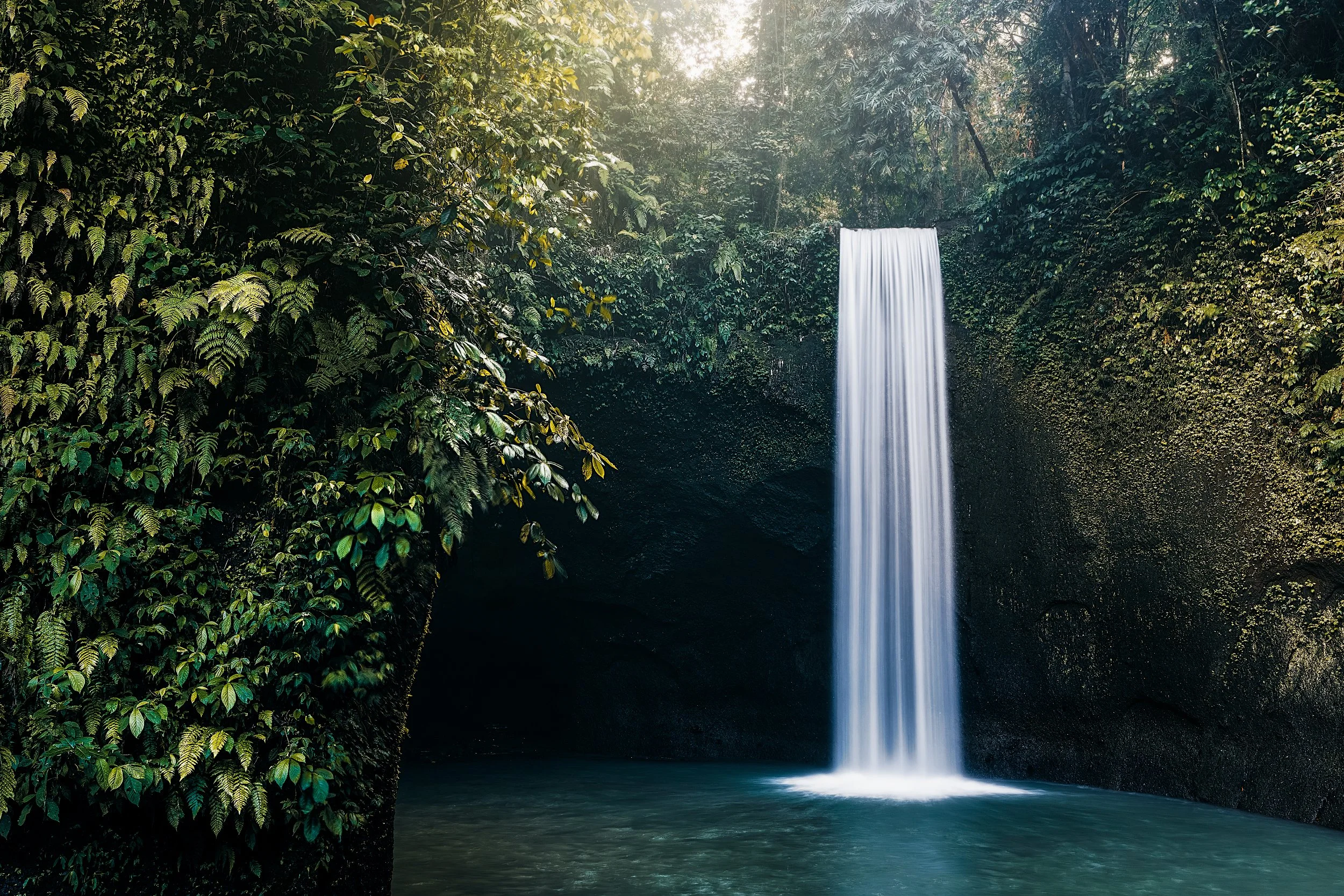 A tall waterfall surrounded by lush green tropical plants and trees, with sunlight filtering through the foliage.