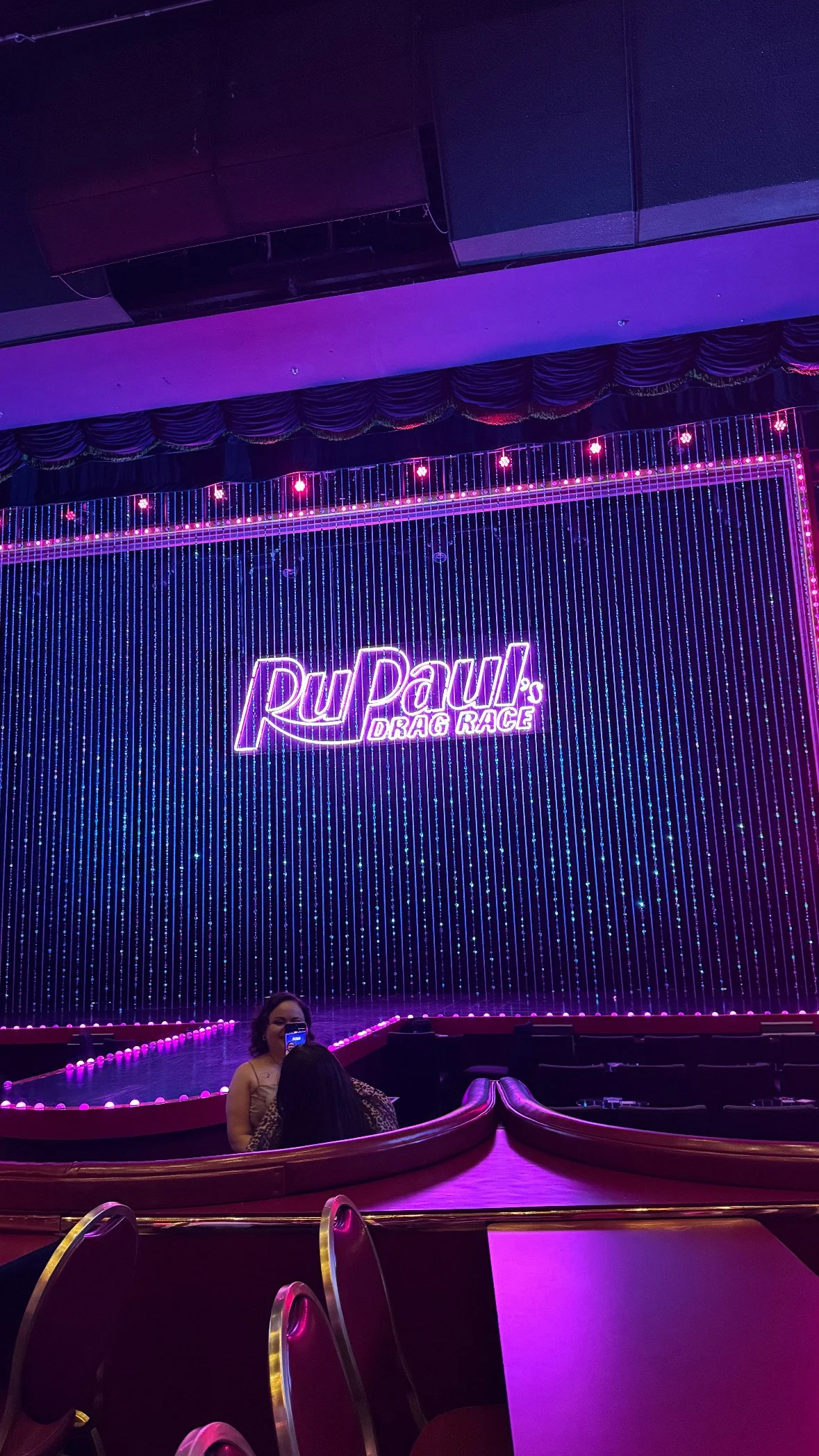 The stage for RuPaul's Drag Race Las Vegas with purple neon sign and curtain of LED lights, and two women sitting in the audience in a theater with velvet seats.