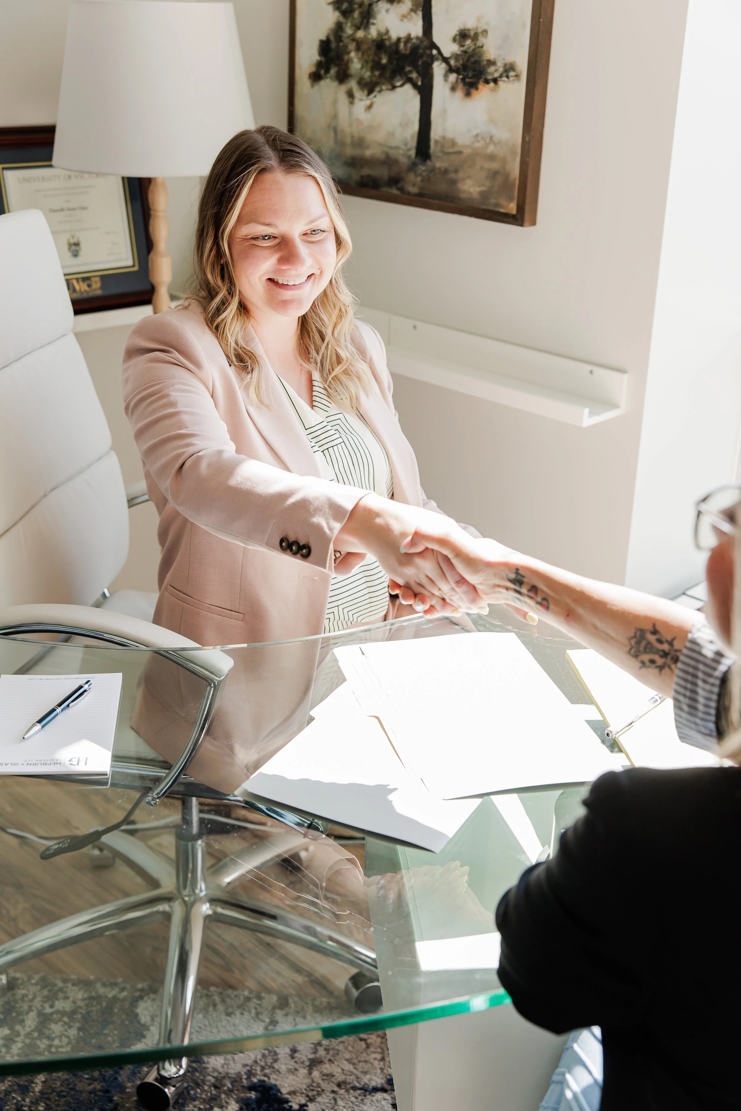 Two women are shaking hands in a professional setting, with one woman seated at a glass desk and the other standing. The seated woman is smiling and wearing a light pink blazer, while the standing woman has tattoos on her arm and wears glasses.