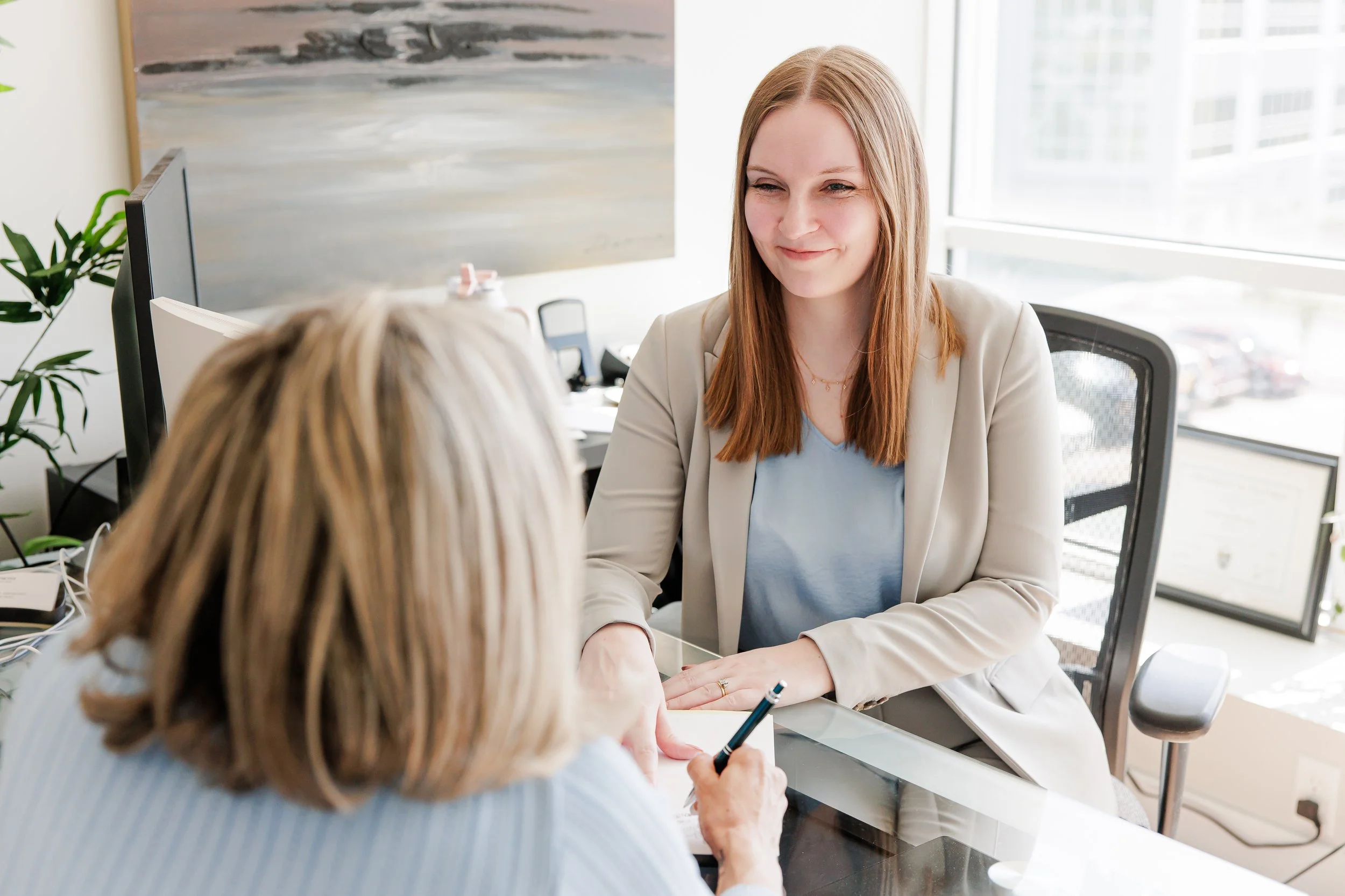 Celeste Cuthbertson family lawyer. Two women having a conversation at an office desk, one with red hair and a white blazer, the other with blonde hair, glasses, and a blue shirt, in a modern office with a large window and computer monitors.