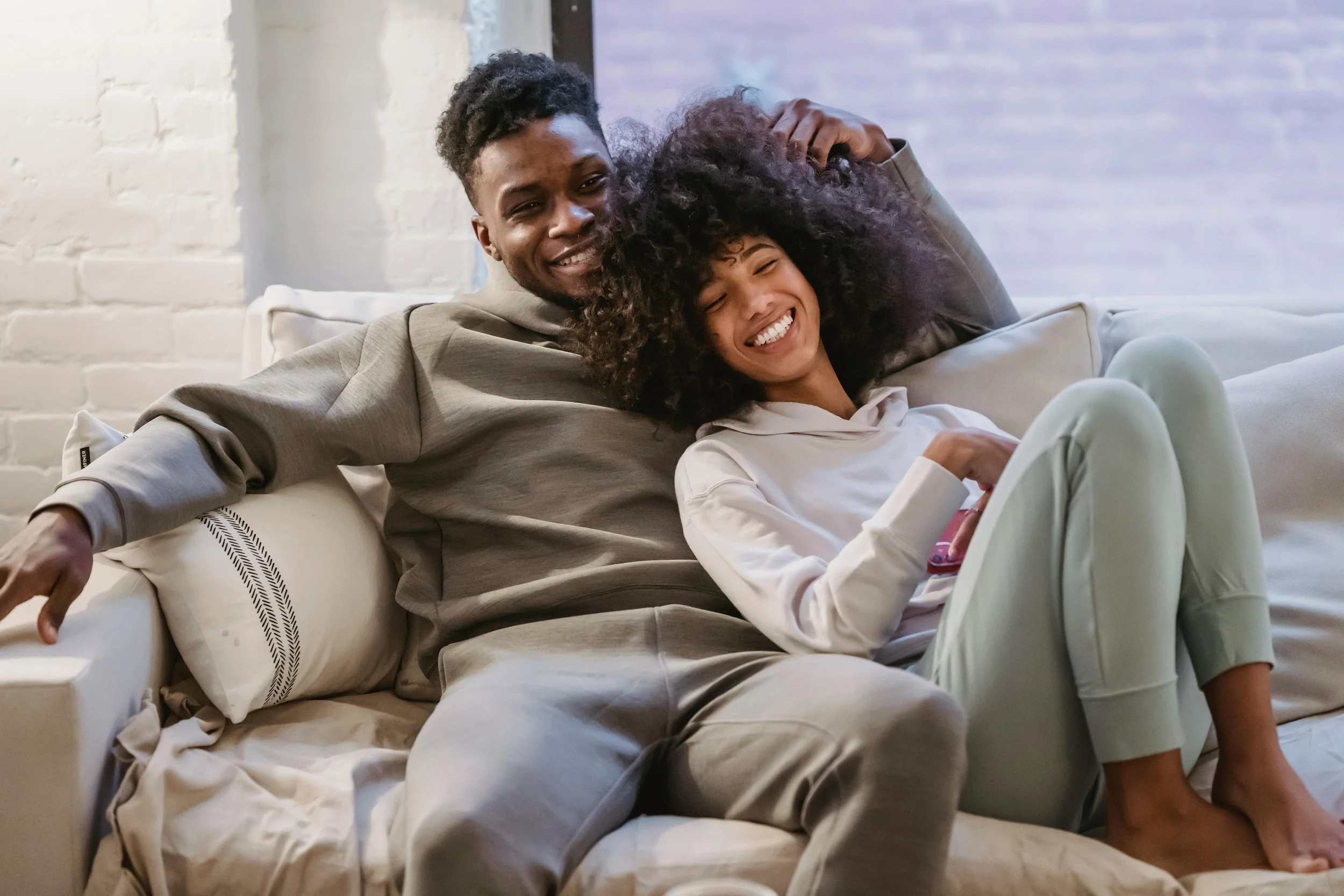 A smiling young man and woman sitting on a couch, with the man's arm around the woman's head, enjoying each other's company in a cozy home setting.