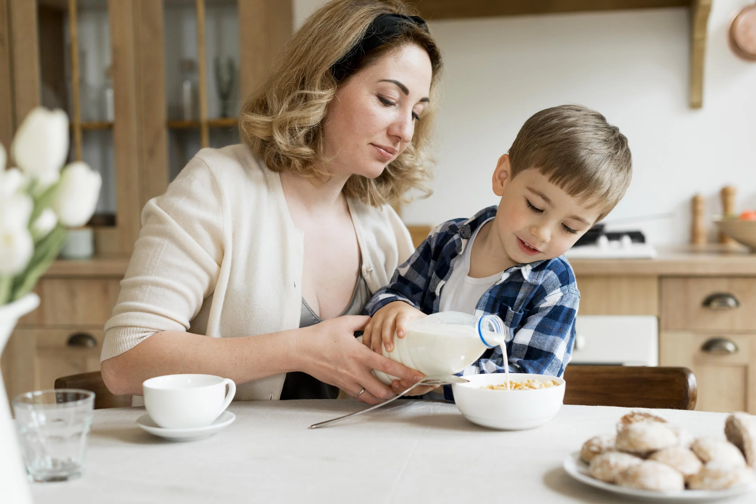A woman and a young boy sitting at a dining table, with the woman pouring milk into a bowl of cereal for the boy in a bright kitchen.