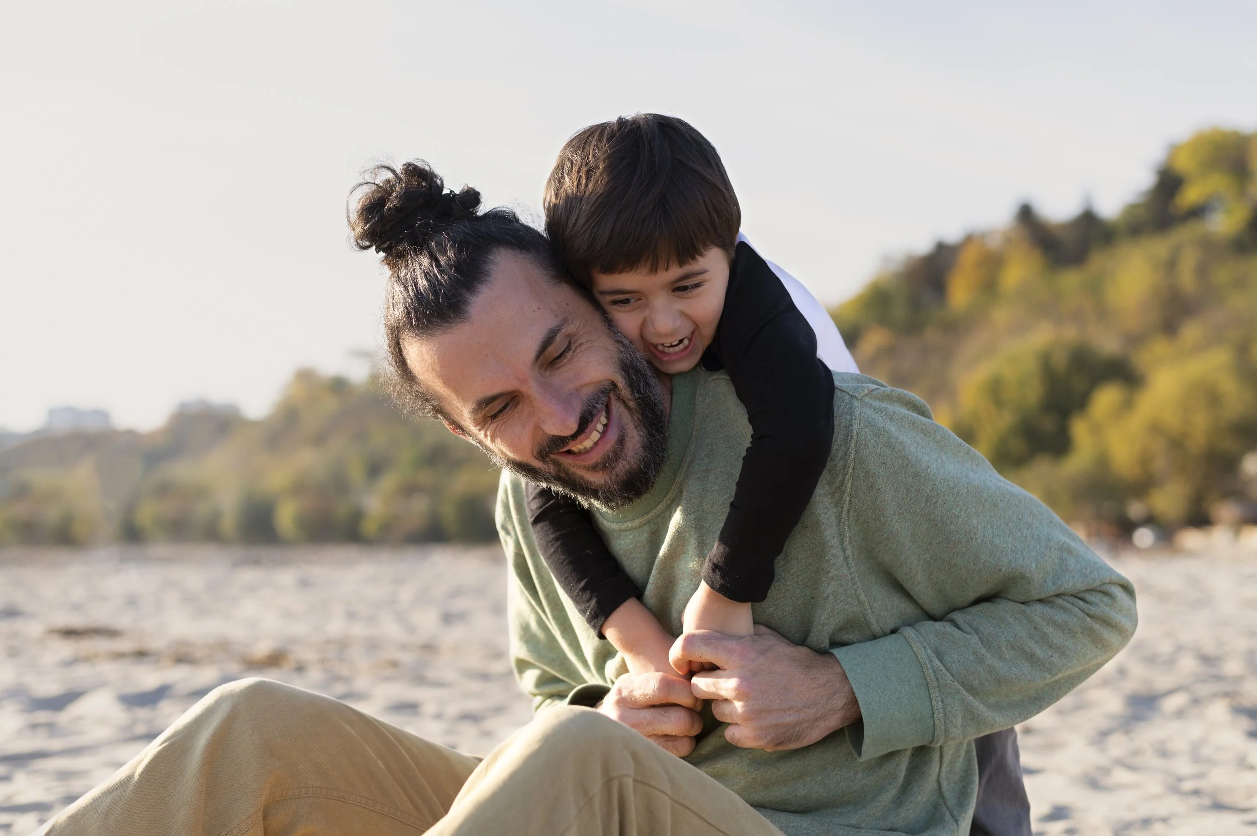 A man with a beard and a bun hairstyle smiling as a young boy with dark hair playfully rides on his back on a sandy beach during daytime, with trees and a clear sky in the background.