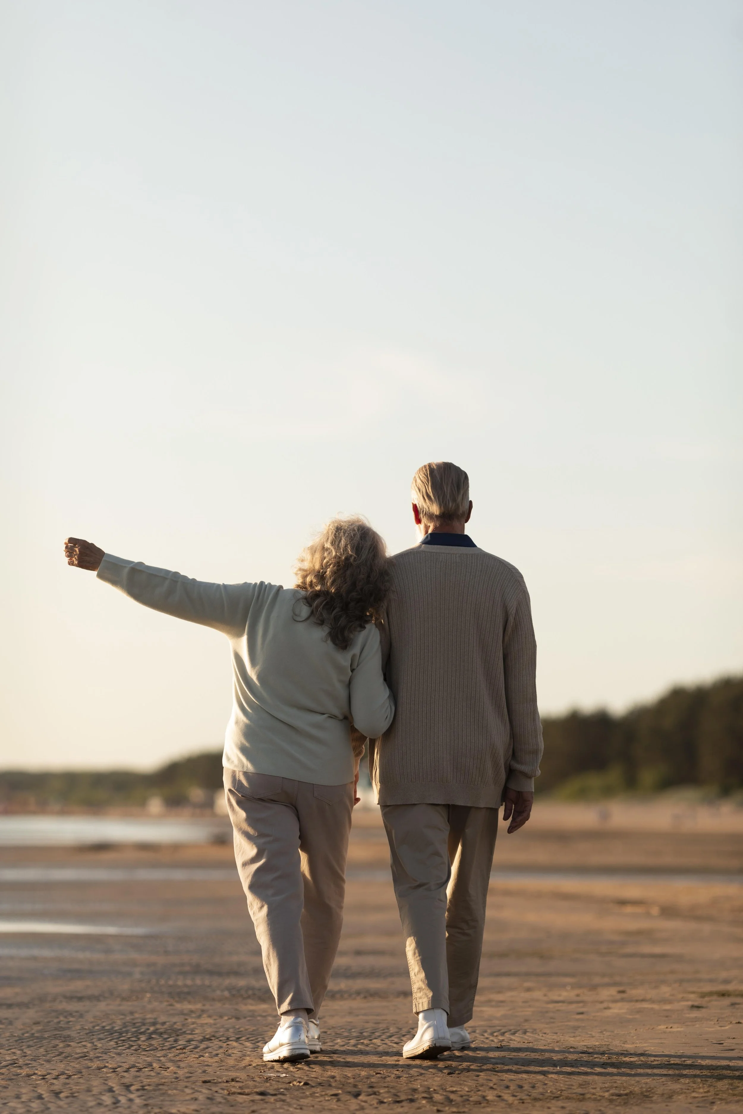 An elderly couple walking together on a beach, with the woman leaning on the man's shoulder and gesturing with her arm, during sunset.