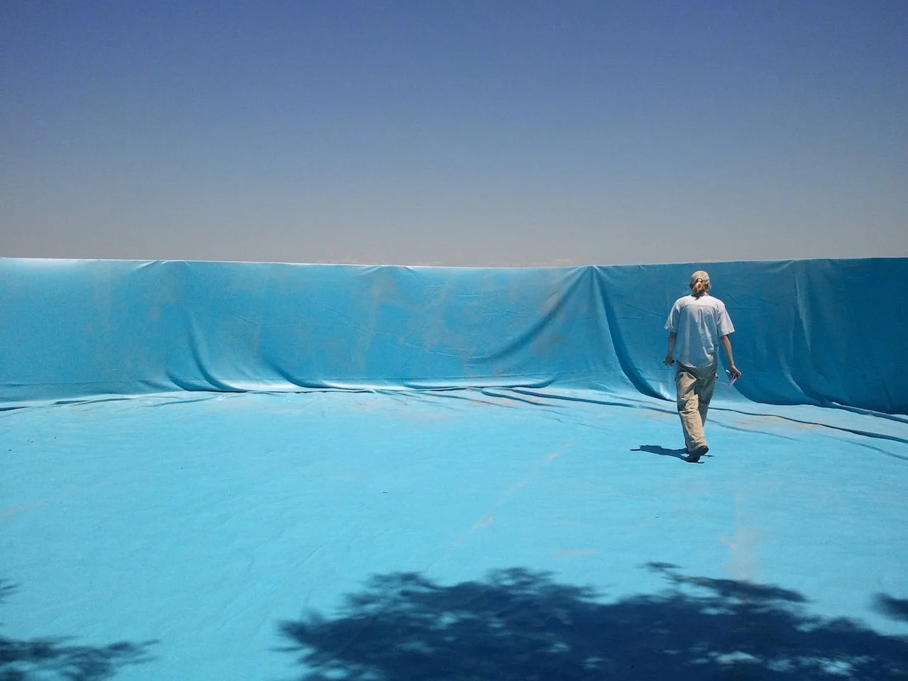 A person walking on a large, flat blue surface with a wall behind them, all covered in a vinyl cover under a clear blue sky. The person's back is to the camera, wearing light-colored pants and a shirt, and holding an object in their right hand.
