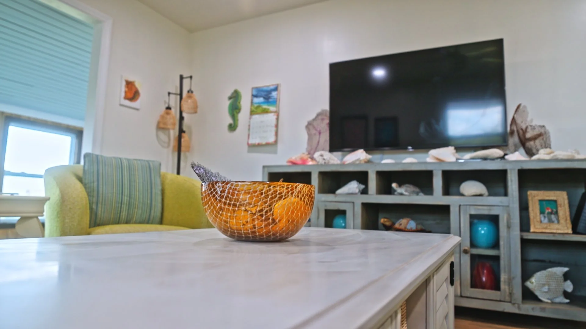Living room with a TV on a cabinet, a yellow and striped armchair, a white table with an orange bowl, and decorative sea shells on the cabinet.