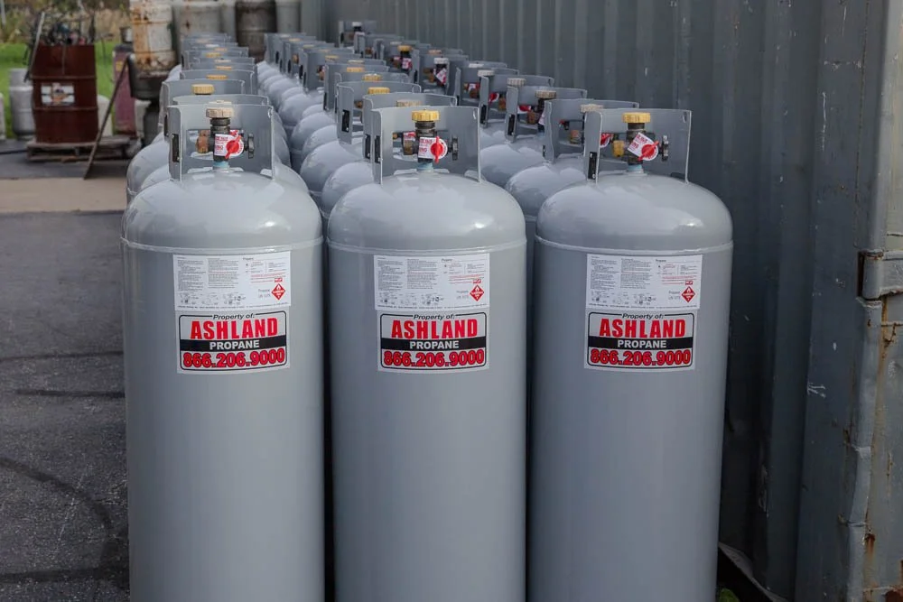Multiple propane tanks labeled with Ashland Propane, arranged in a row outside near a metal storage container and other equipment.
