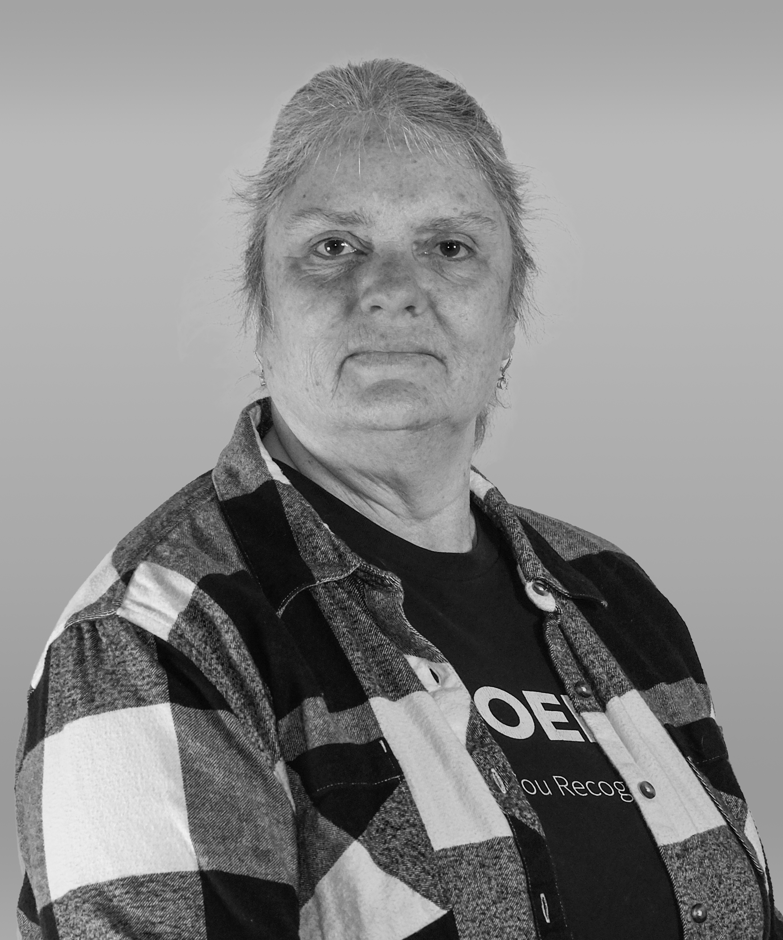 A black-and-white portrait of an older woman with short hair, wearing a plaid shirt over a T-shirt, facing the camera with a neutral expression.