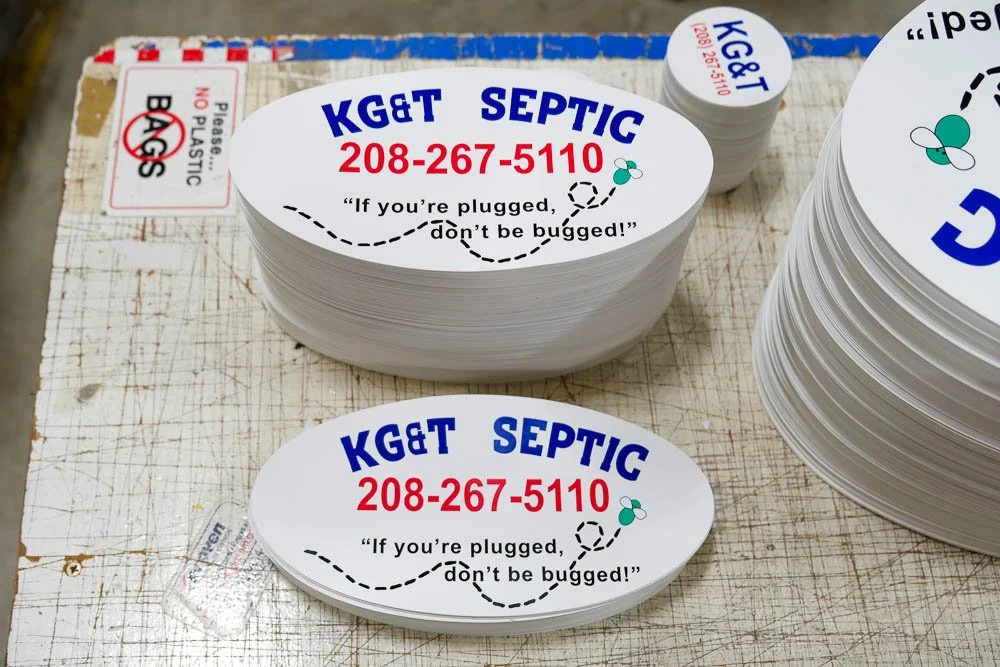 Oval stickers advertising KG&T Septic with a phone number 208-267-5110 and a tagline "If you're plugged, don't be bugged!" in blue and red text, displayed on a work table.
