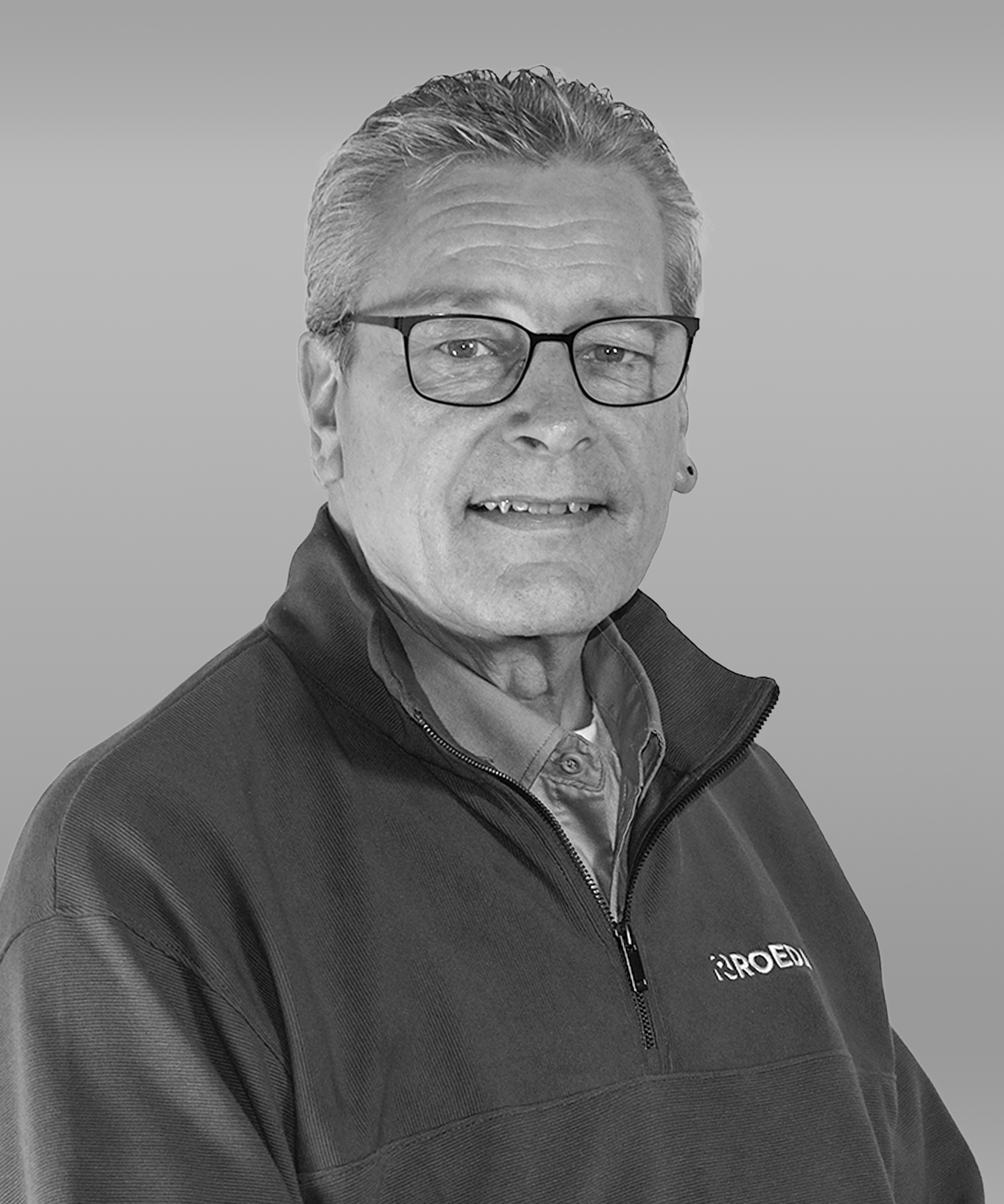 Black and white portrait of an older man with glasses, wearing a zip-up jacket with 'IROEDO' logo, smiling slightly, looking at the camera.