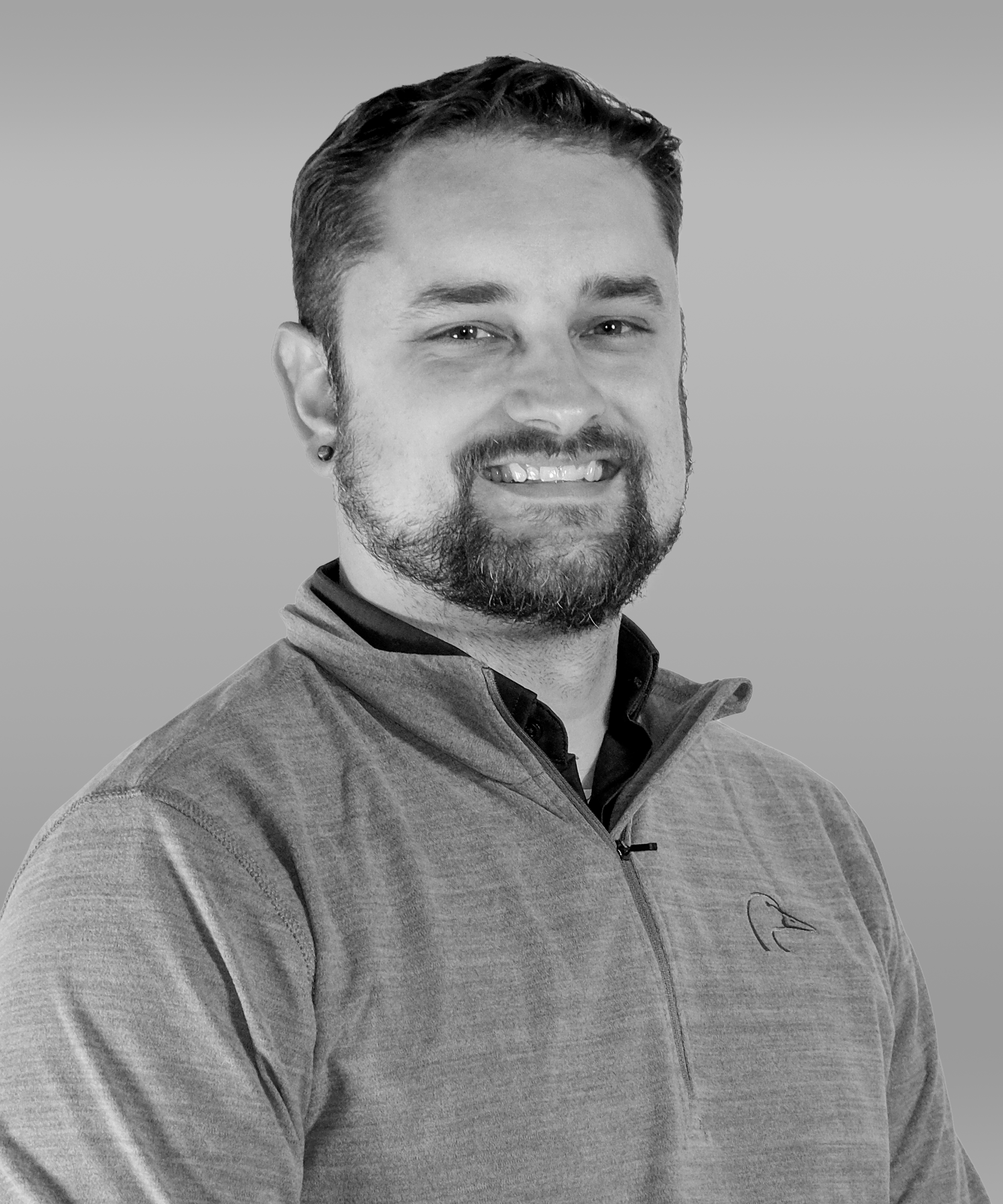 Black and white photo of a smiling man with short hair, a beard, and wearing a quarter-zip jacket with a logo on the chest.