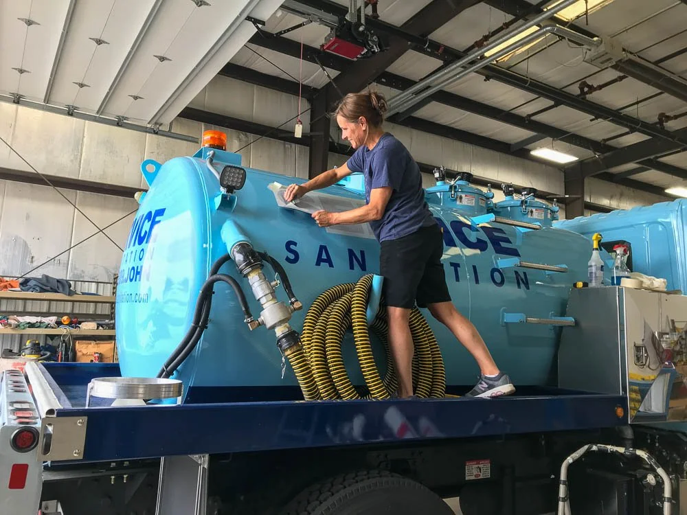 A woman cleaning a large blue liquid tank labeled 'SANITATION' in an industrial warehouse.