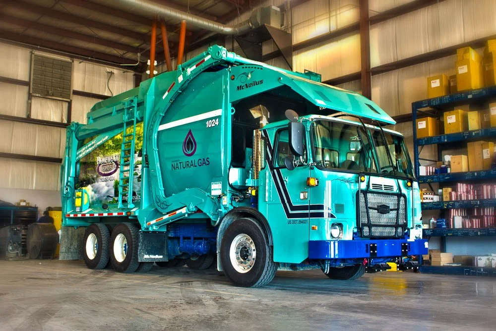 A teal-colored garbage truck with 'NATURALGAS' branding is parked inside a warehouse. The truck has a prominent front grille, multiple wheels, and a rear loading area.