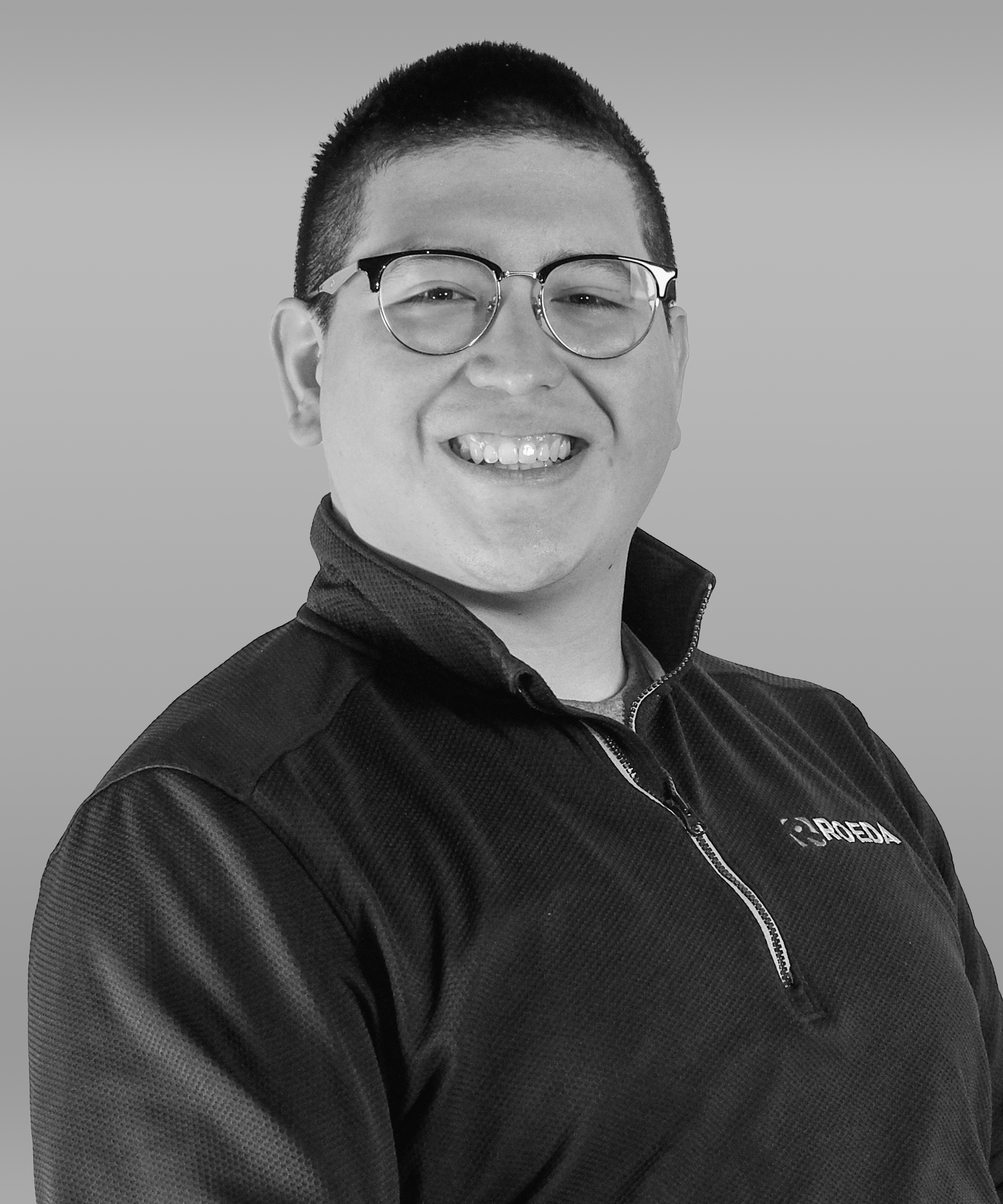 Black and white portrait of a smiling man wearing glasses and a quarter-zip athletic jacket with a logo on the chest, against a plain background.