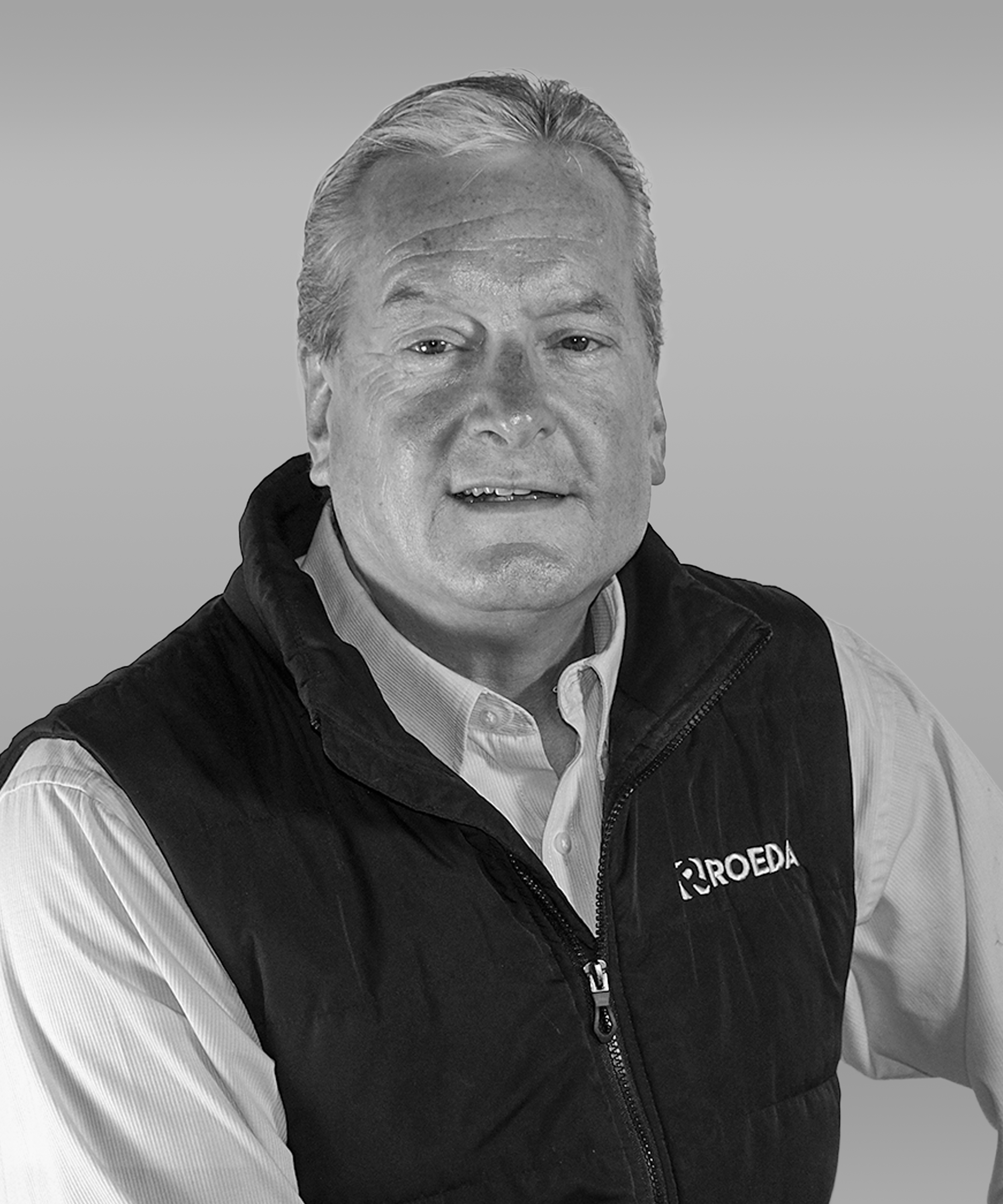 Black and white photo of a middle-aged man with light-colored hair, wearing a white collared shirt and a black vest with a logo, against a plain background.