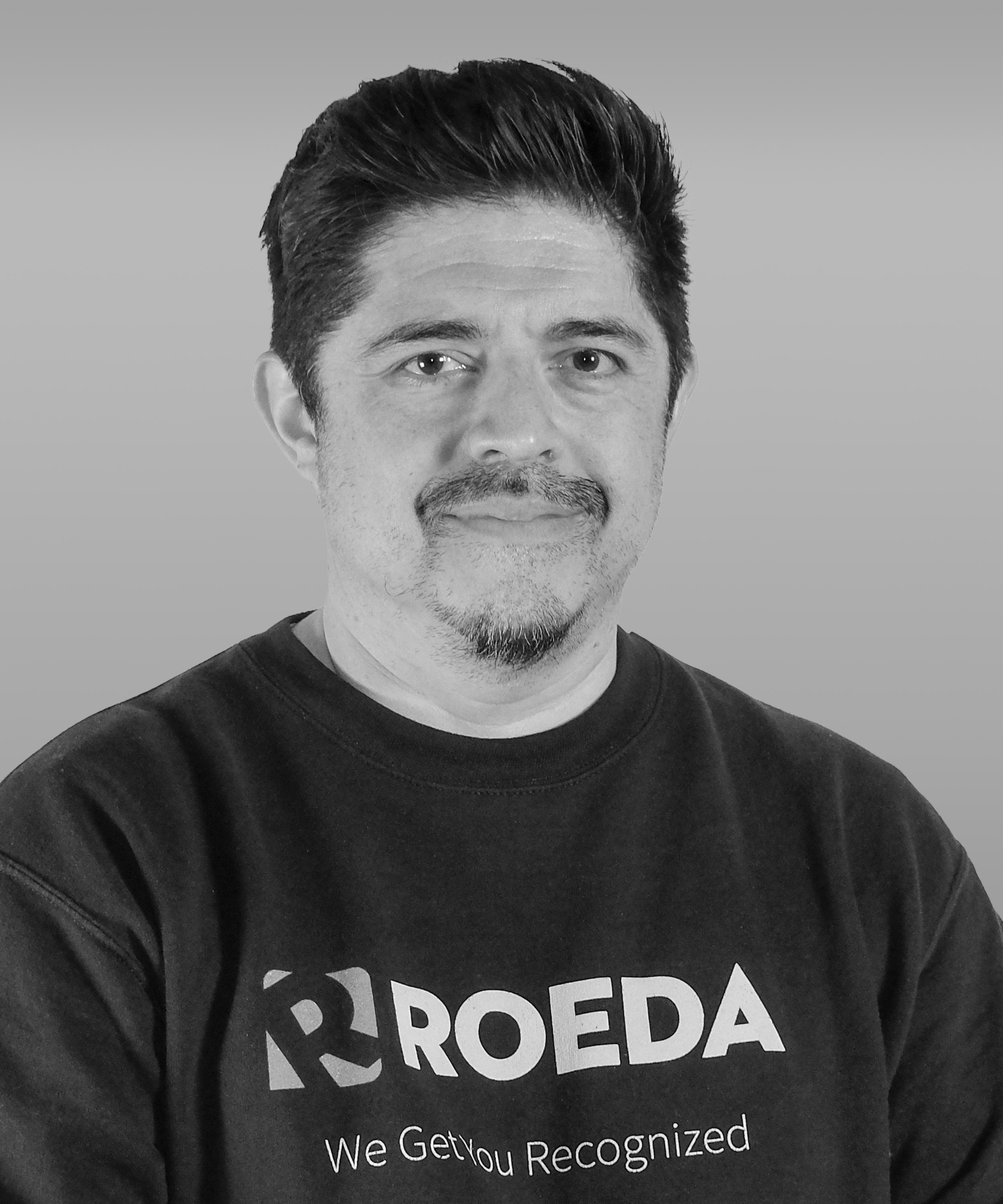 Black and white photo of a man with short dark hair, facial hair, wearing a T-shirt with the logo 'ROEDA' and the slogan 'We Get You Recognized'.