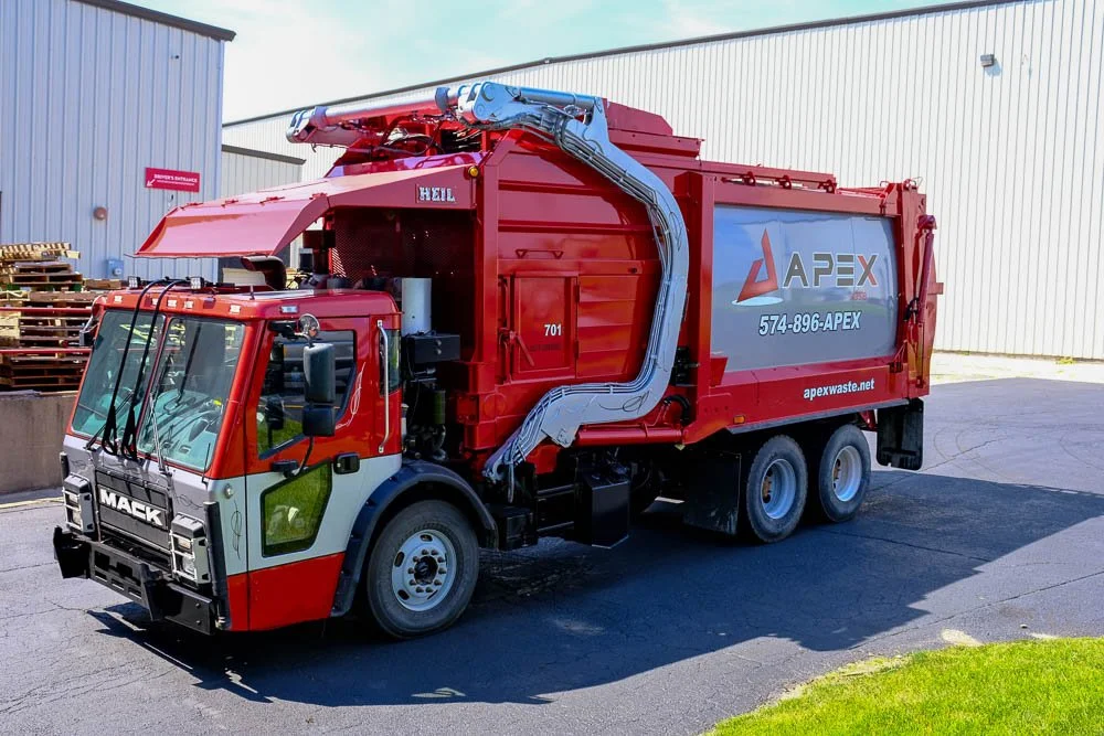 Red and white Mack garbage truck with Apex Waste branding parked outdoors near a warehouse