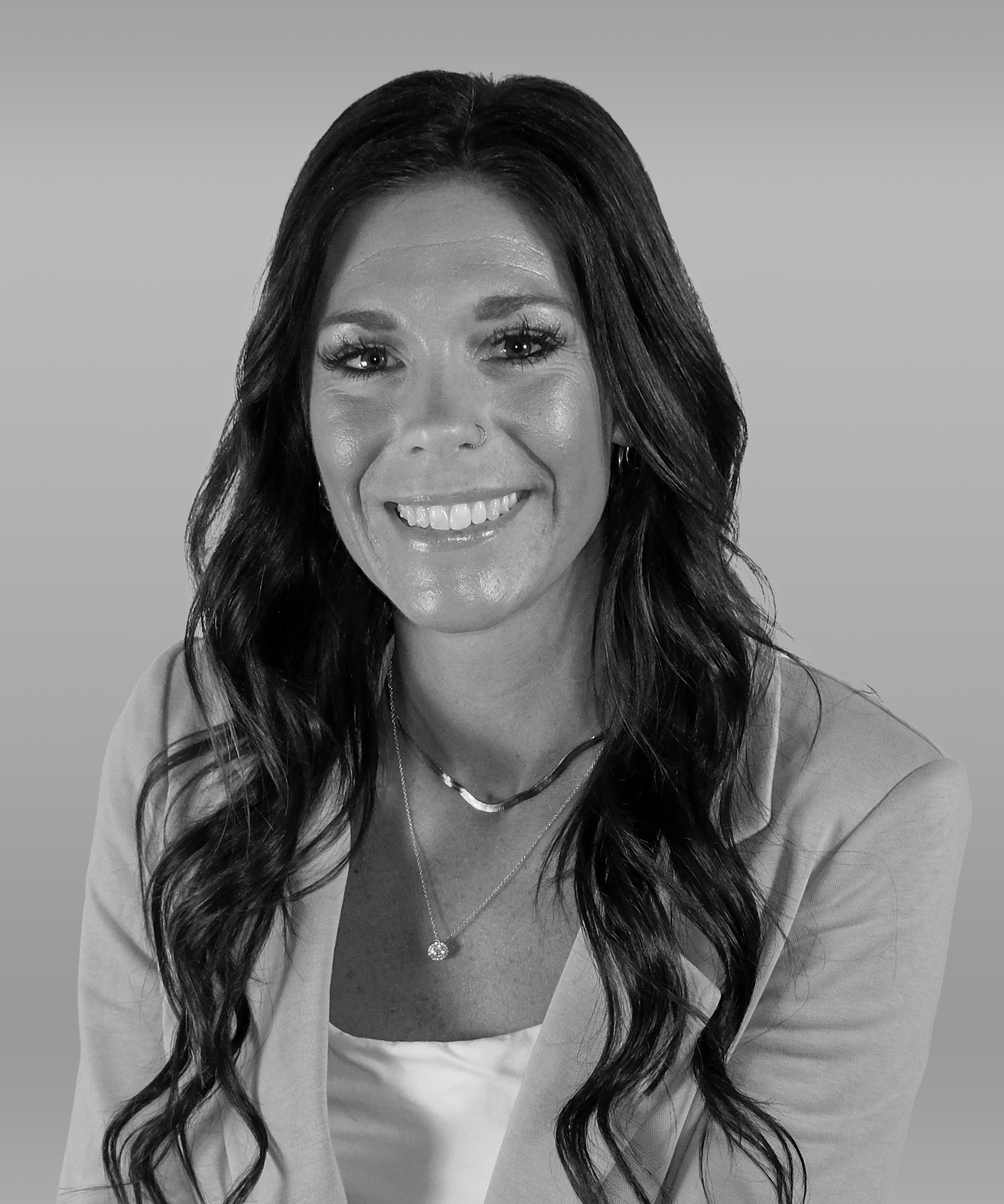 Black and white portrait of a smiling woman with long wavy dark hair, wearing a light-colored blazer, a necklace, and earrings, against a plain background.