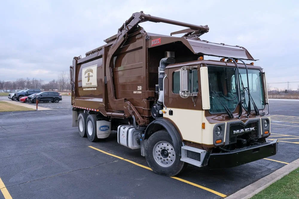 A brown and cream-colored Mack front-loading trash truck parked in a parking lot.