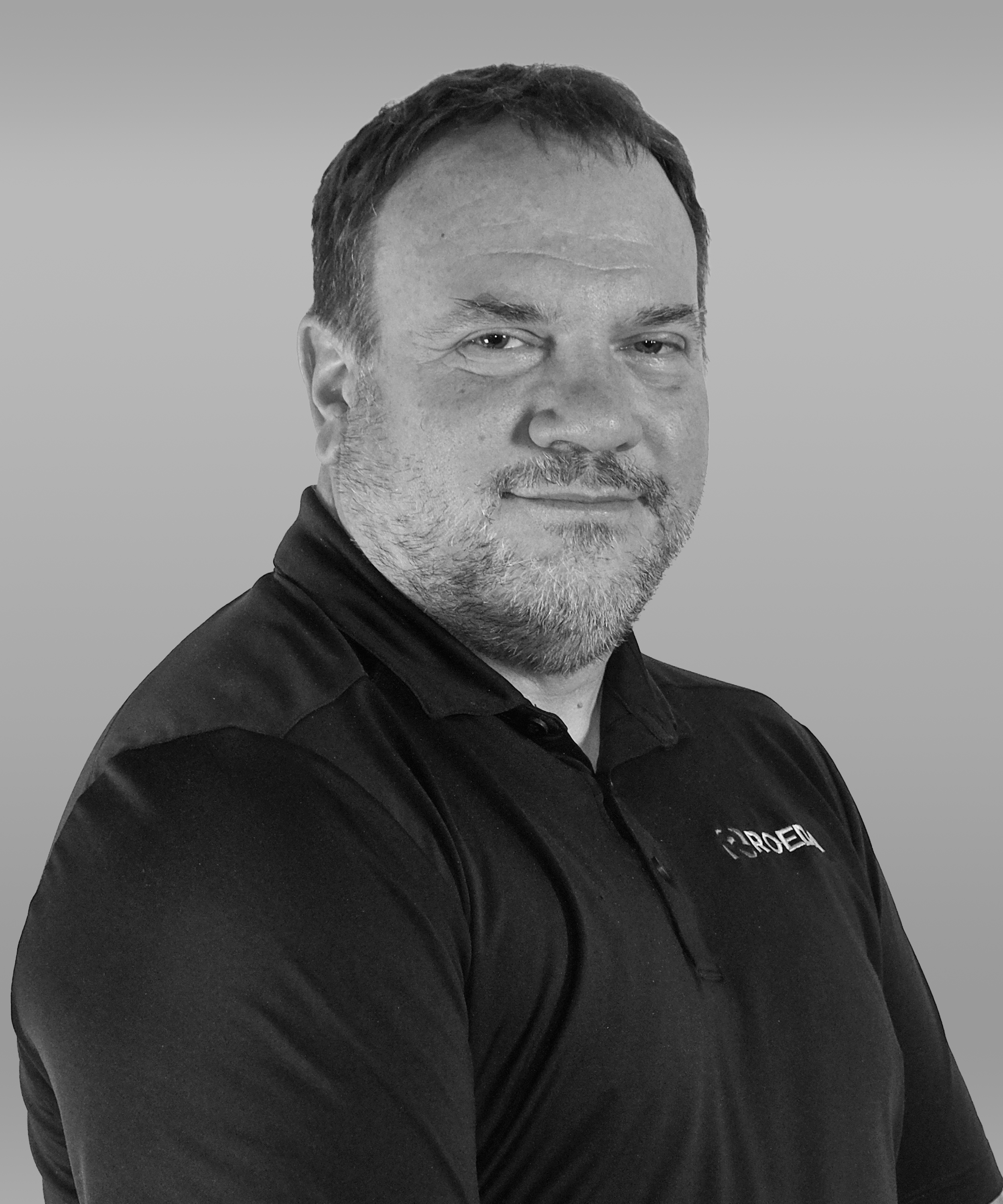 Black-and-white portrait of a man with short hair and a beard, wearing a collared shirt with a logo, against a plain background.