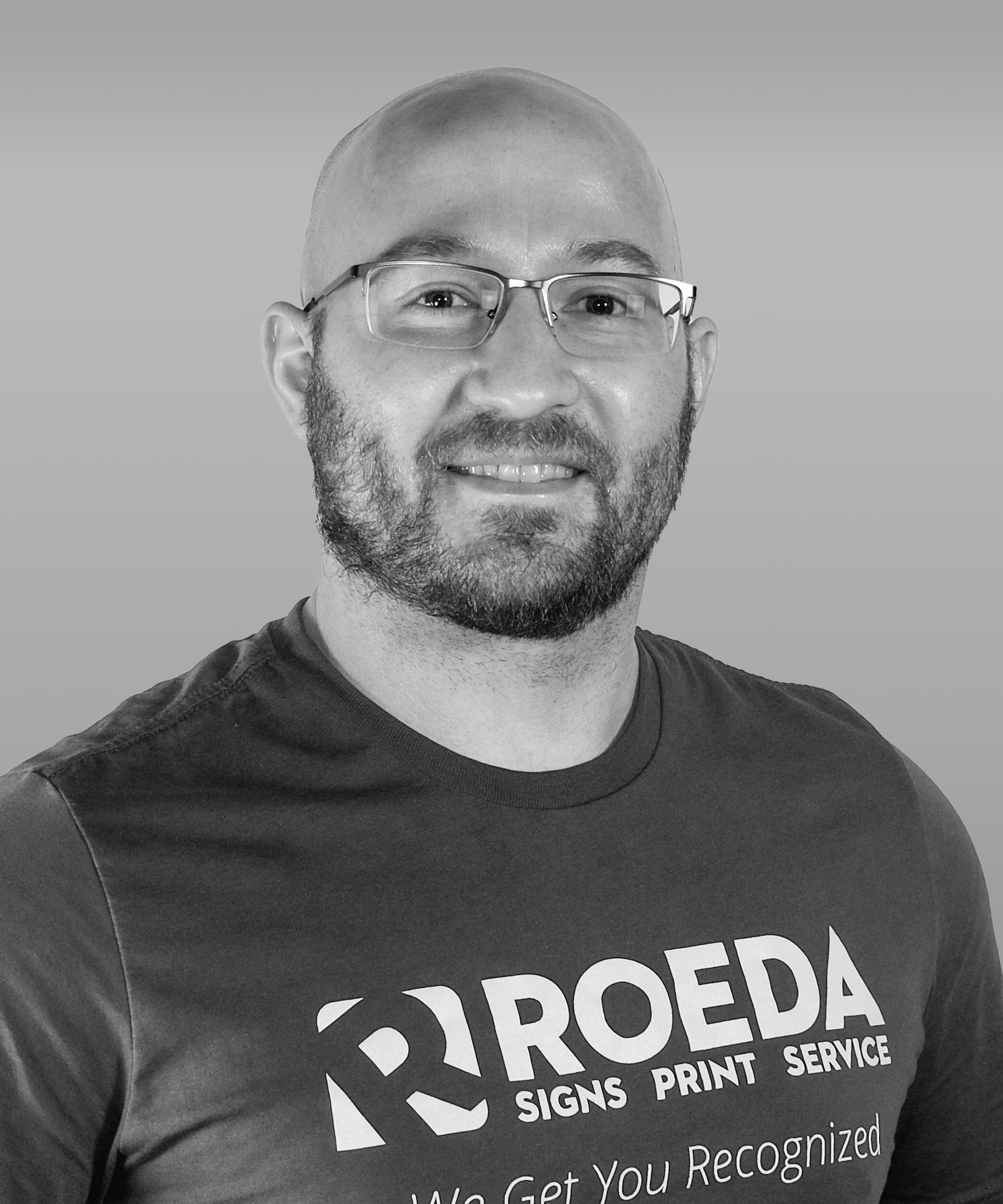 Black and white photo of a man with glasses, a beard, and a shaved head, wearing a t-shirt with the logo and text 'ROEDA Signs Print Service, We Get You Recognized'.