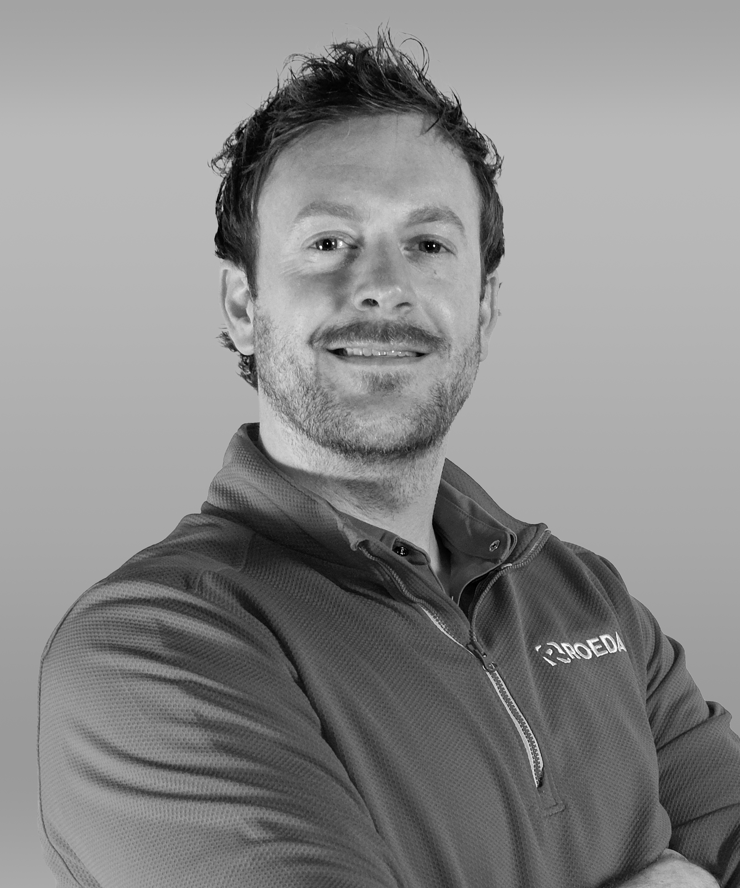 A man with dark, wavy hair and a light beard, wearing a branded jacket, smiling with arms crossed against a plain background.