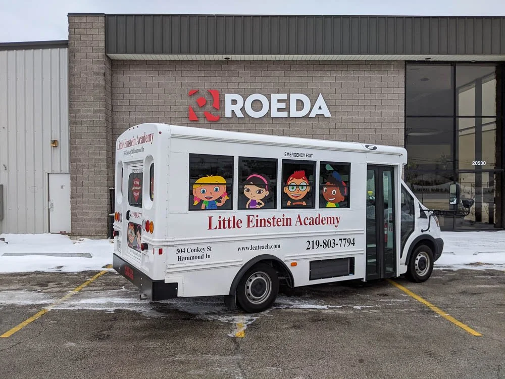 A small bus with cartoon children and the text 'Little Einstein Academy' parked in front of a building with a 'ROEDA' sign.