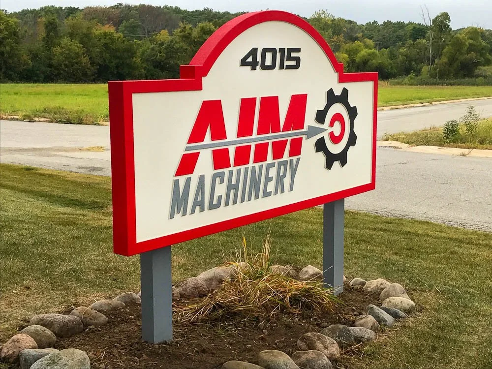 Business sign for AIM Machinery with the street number 4015 in a rural area with grass, trees, and a sidewalk.