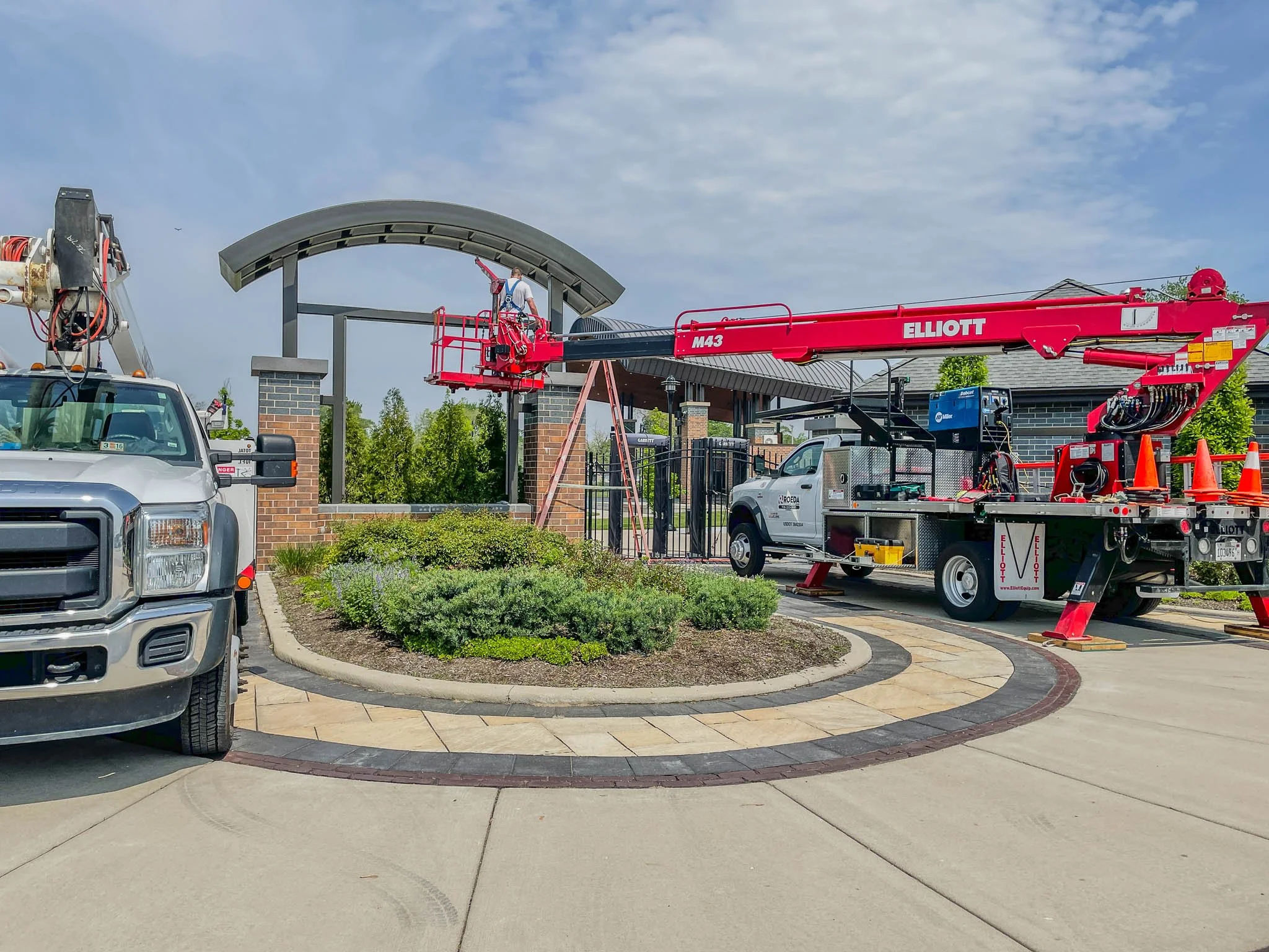 Construction workers using a cherry picker lift to work on a brick entrance gate with a curved roof, surrounded by landscaping and parked trucks.