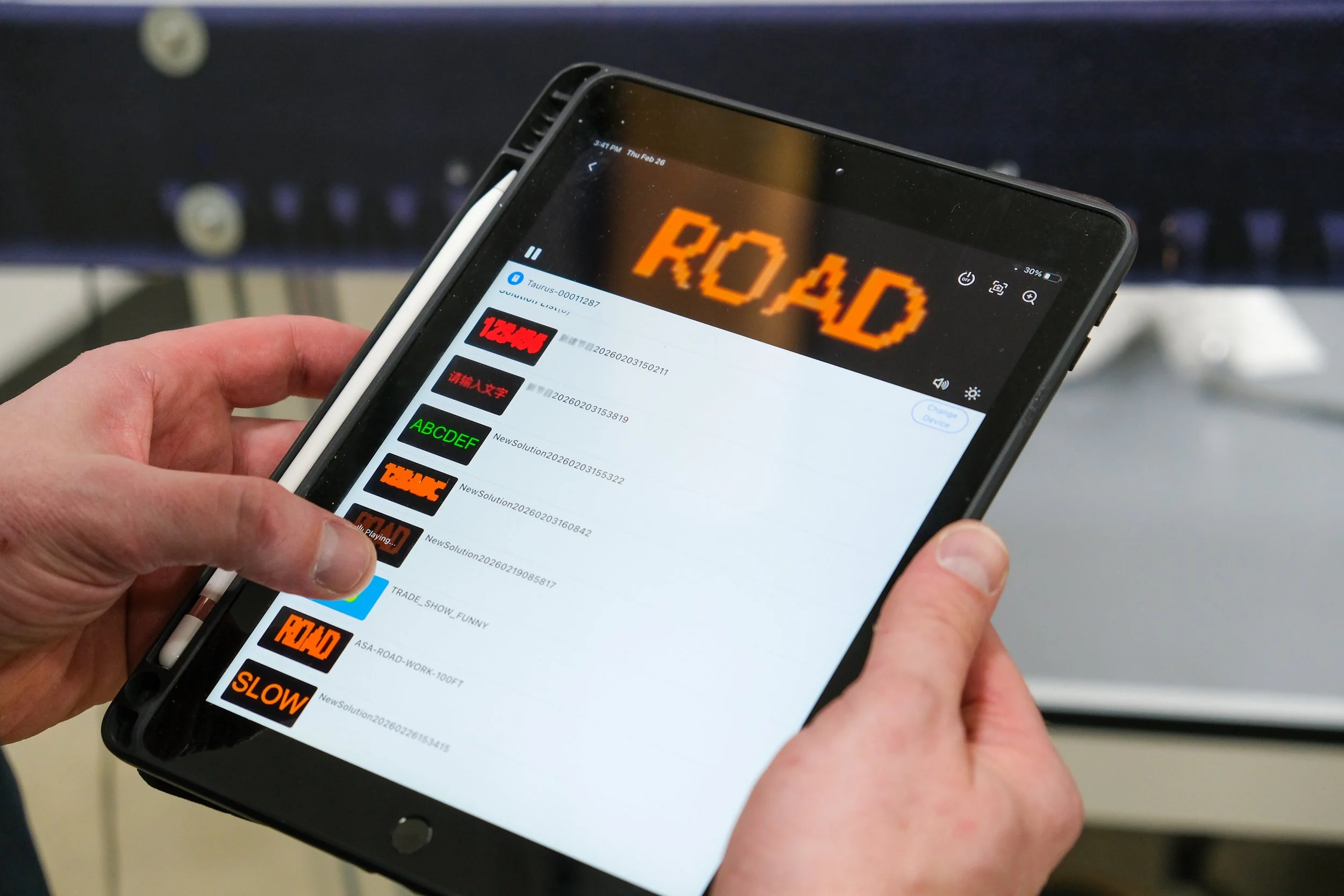 A person holding a tablet shows a screen with various video thumbnails and the word ‘ROAD’ in orange pixelated text on a separate device in the background.
