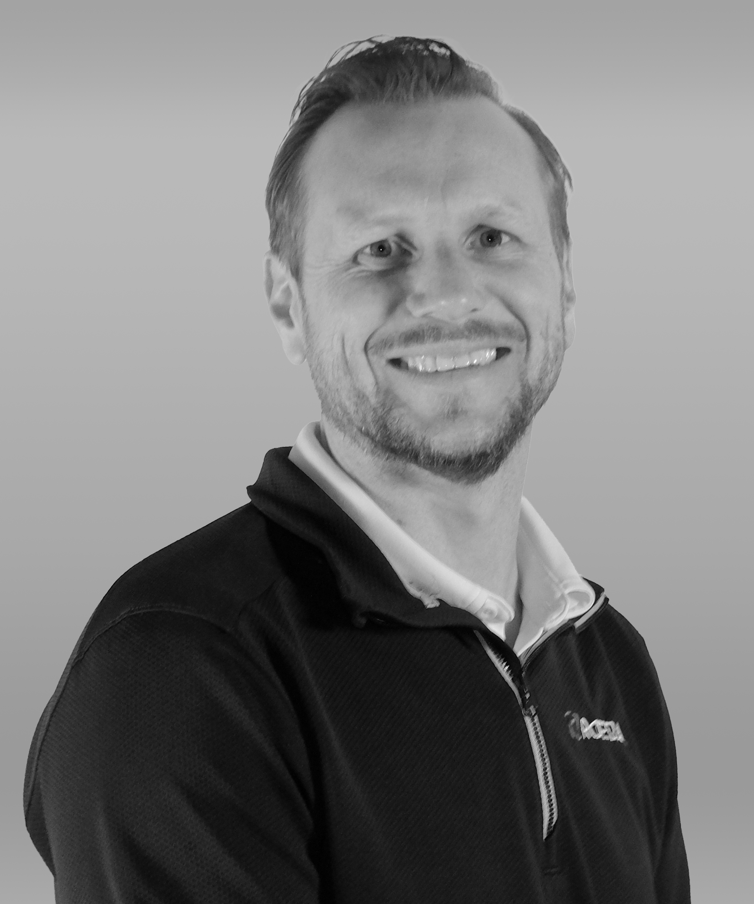 Portrait of a smiling man with light-colored hair and beard, wearing a black jacket over a white collared shirt, against a plain background.