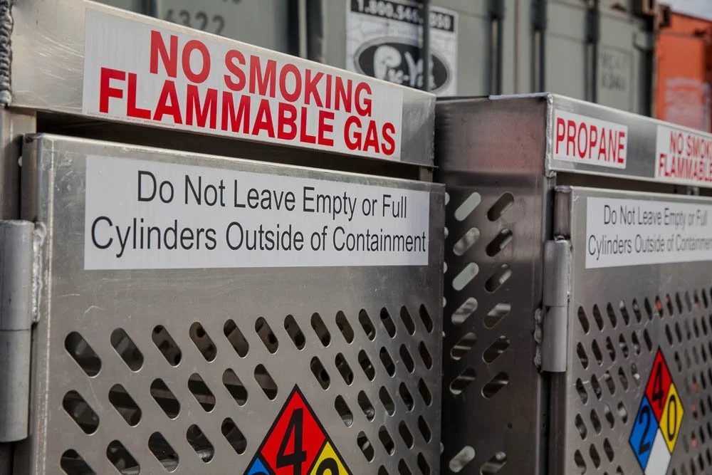 Metal gas cylinders storage units with warning signs that read 'No Smoking Flammable Gas,' 'Propane,' and instructions to not leave empty or full cylinders outside of the containers.