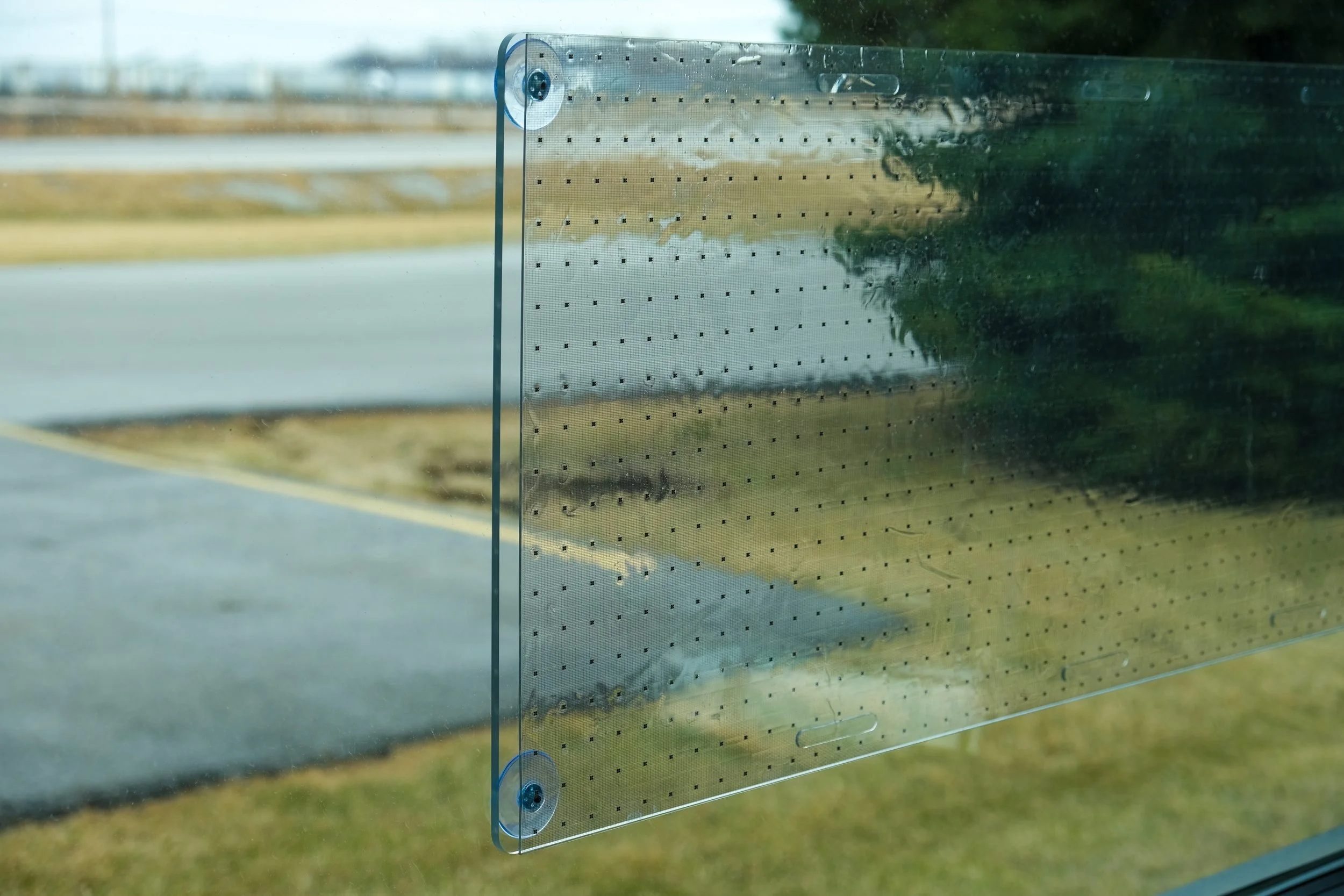 A clear glass or acrylic panel with suction cups mounted on a window, showing a suburban street with grass, trees, and a bridge in the background.
