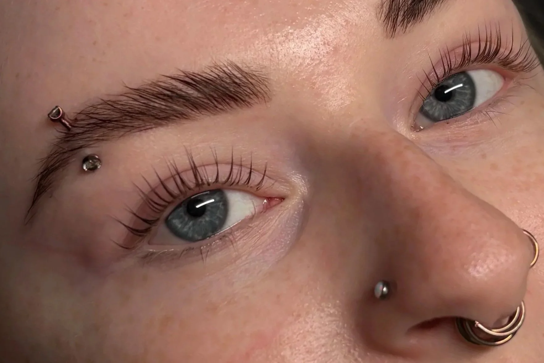 Close-up of a person's face focusing on both eyes, showing well-groomed eyebrows with embedded jewelry, long eyelashes, and blue eyes with visible irises and pupils.