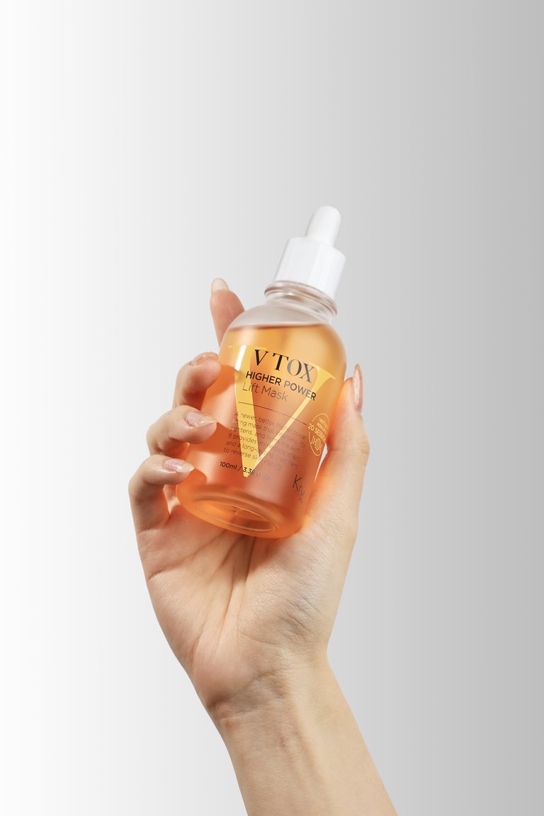 Close-up of a hand holding a small bottle of orange liquid serum or facial treatment with a white dropper cap, against a plain background.