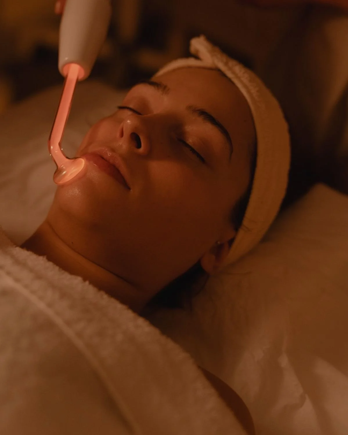 A woman lying down with her eyes closed, receiving a facial treatment using a pink facial tool on her lips.