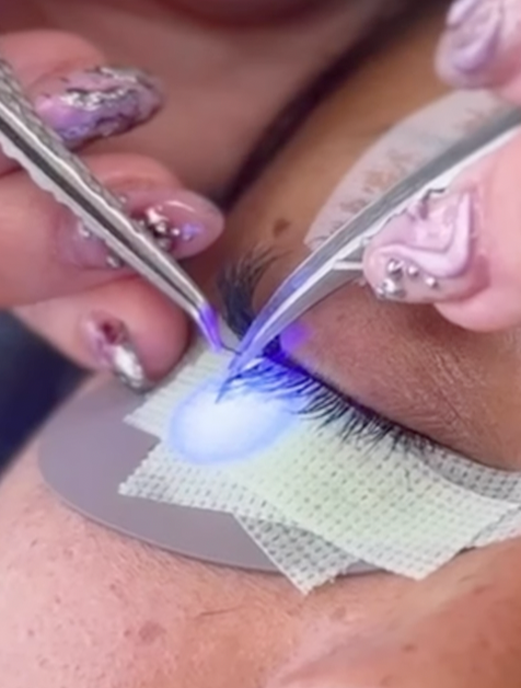 A close-up of a person undergoing an eyelash extension procedure, with two technicians applying eyelash extensions using tweezers and a blue light to cure the adhesive, while wearing nail polish with rhinestones.