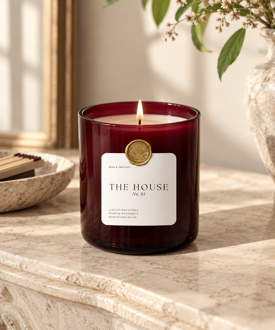 A lit red candle in a glass jar with a label that reads "The House No. 01" sitting on a marble surface, with a decorative bowl and a vase with flowers in the background.