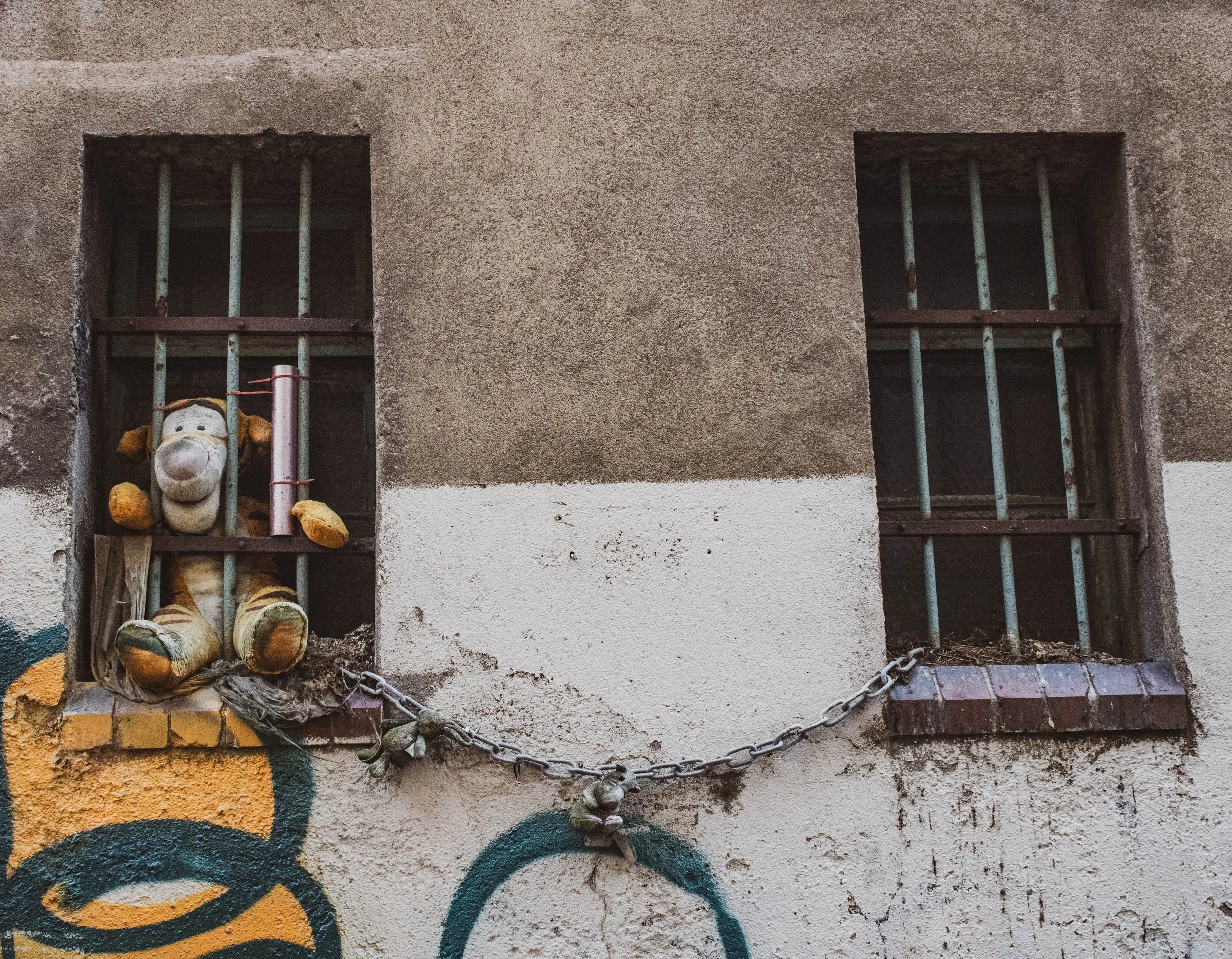 A worn-out stuffed animal in a jail cell window with rusty bars, chained to a wall with graffiti.