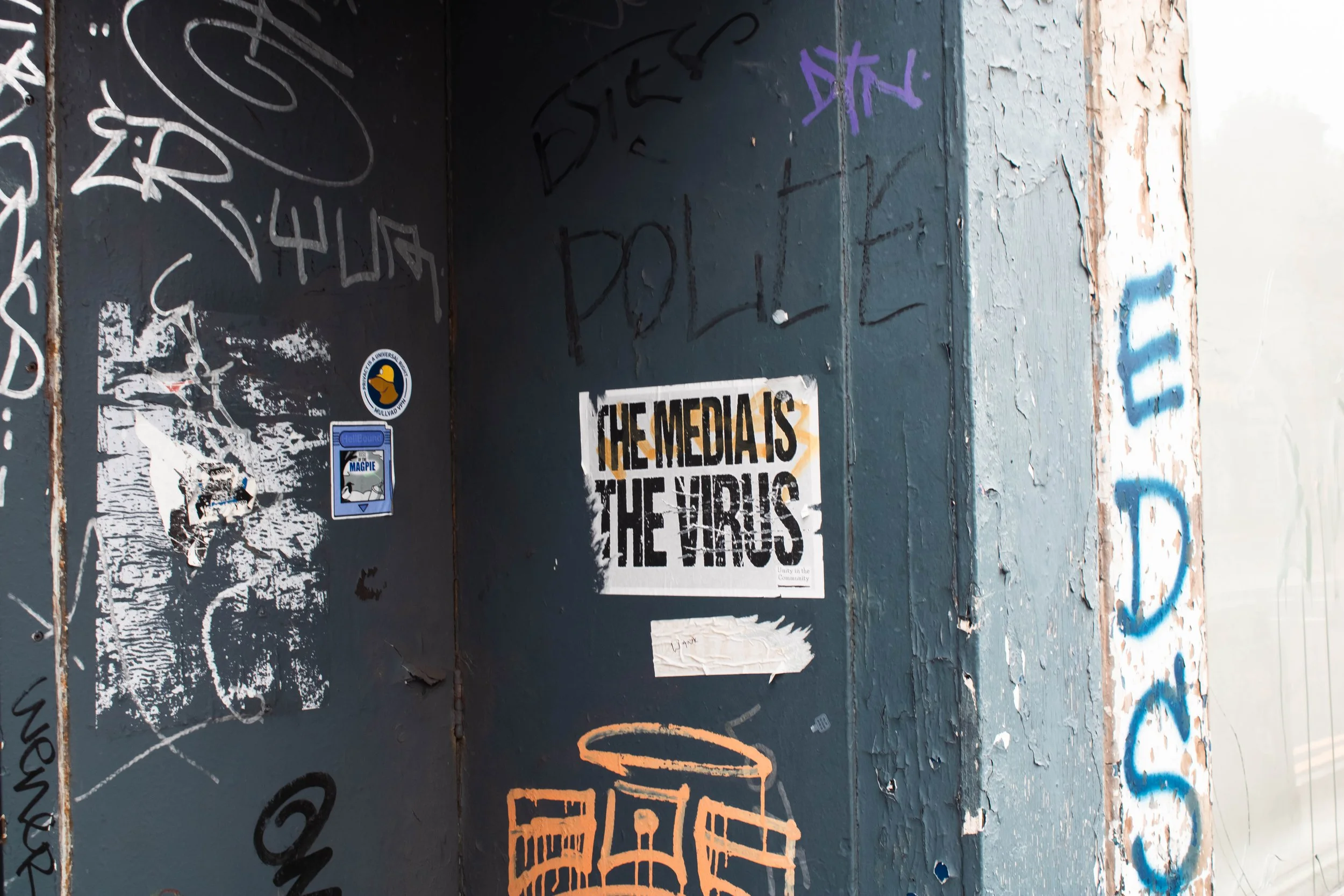 A wall with graffiti and stickers, including a sticker featuring a duck, a sticker with the word 'MAGPIE,' and a printed poster that reads 'THE MEDIA IS THE VIRUS.' The wall is peeling and weathered, with various scribbles and tags.