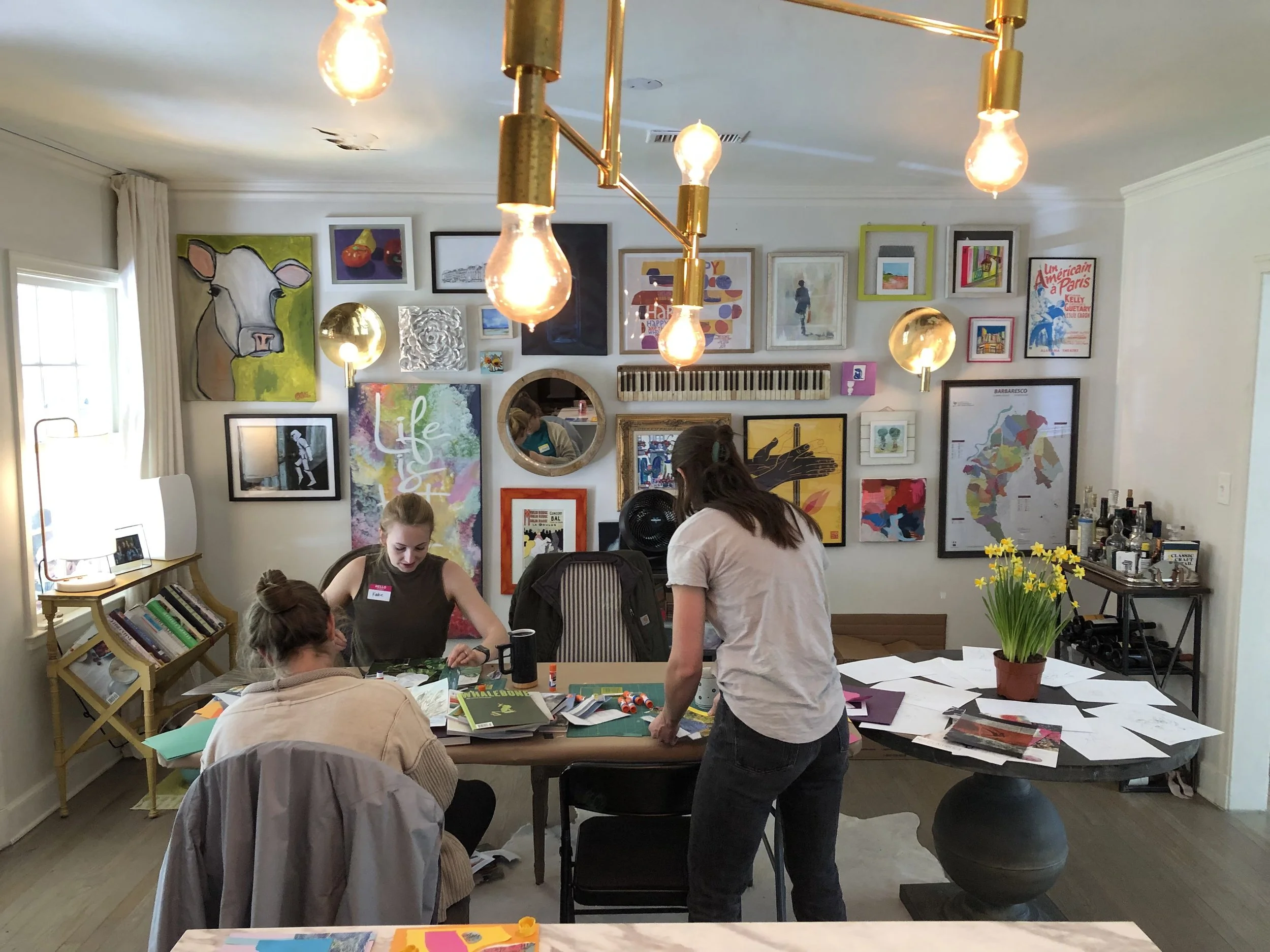 A group of three women working and crafting at a cluttered table in an art-filled room with a gallery wall of colorful artworks and photographs, some on note papers and scissors, with bottles of liquor on a cart in the background.