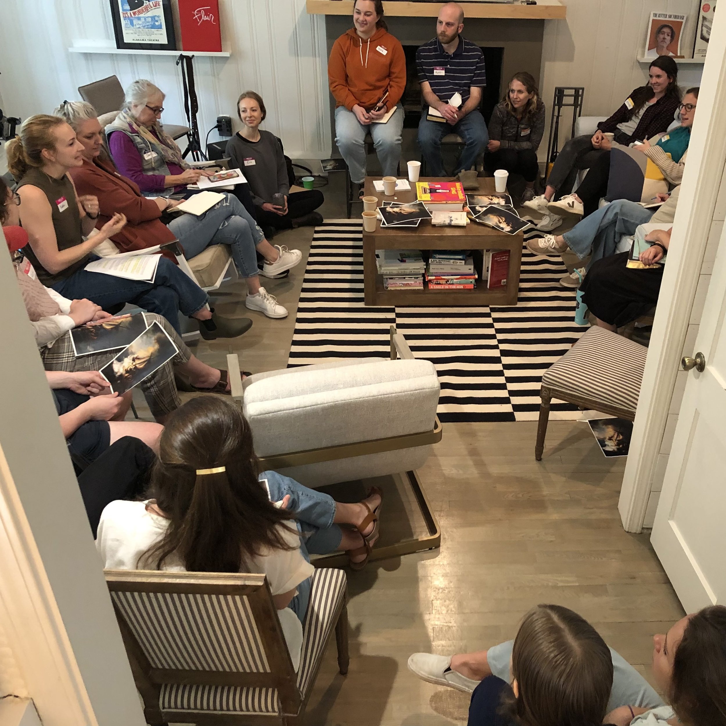A group of people sitting in a cozy room, engaged in a discussion or meeting, with some holding photographs and notebooks, and a coffee table in the center with books and cups.