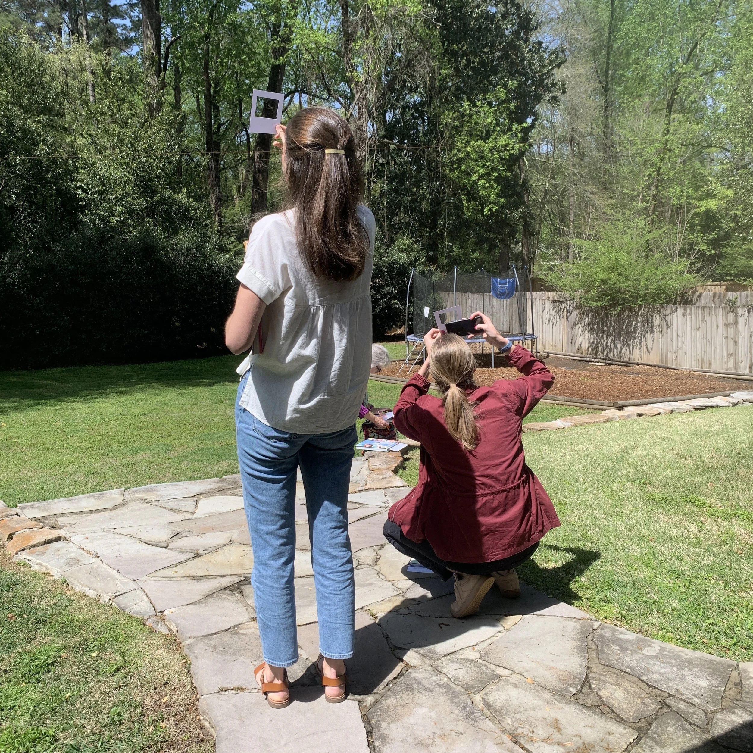 Two women are outside in a backyard, one standing and one kneeling, both taking photos of a framed picture of a square with a circle inside. The yard has green grass, trees, a wooden fence, and a trampoline in the background.
