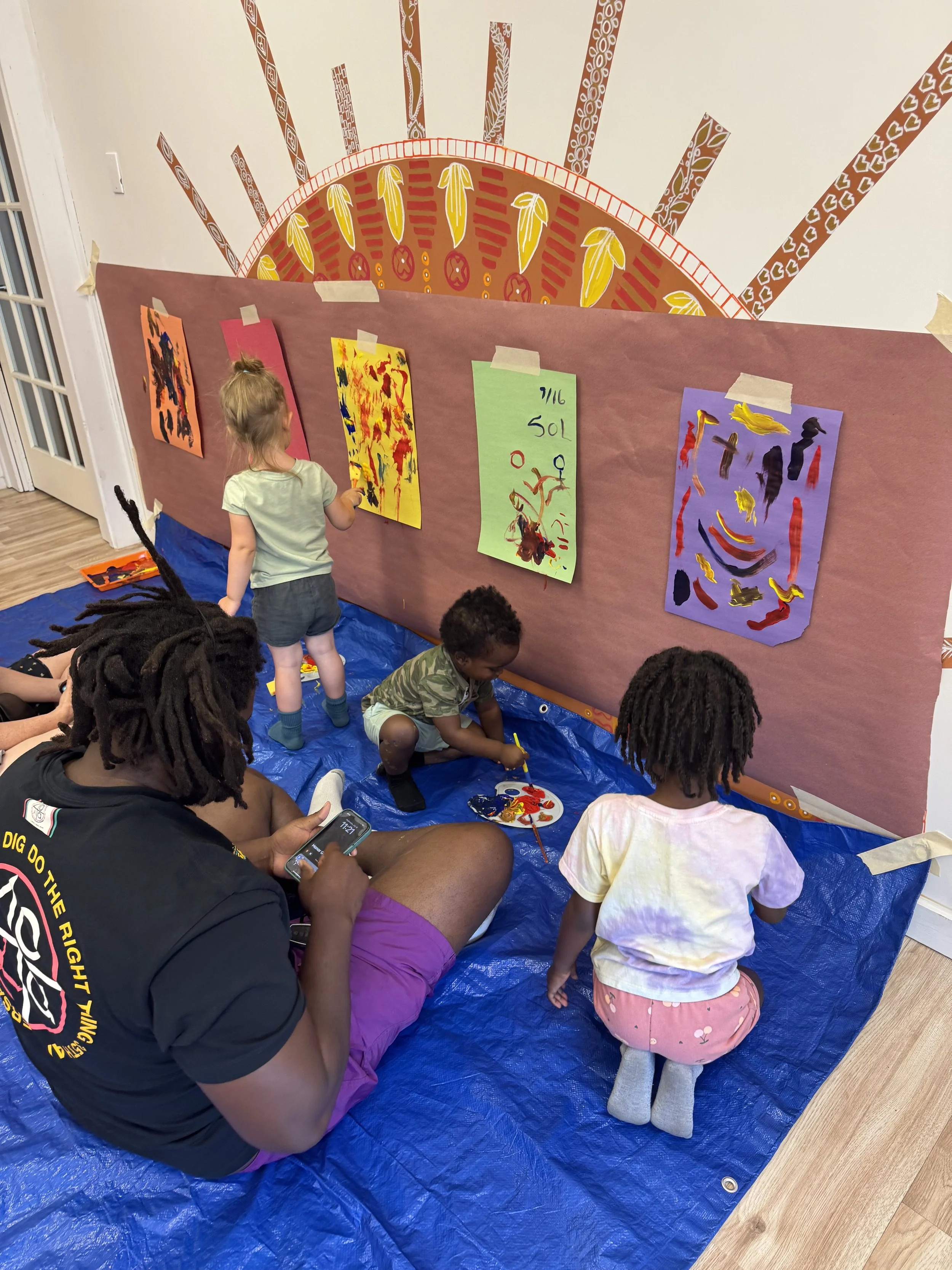 Children and an adult painting and observing artwork on a brown paper-covered wall with colorful abstract paintings, sitting on a blue tarp in a room with wooden flooring.