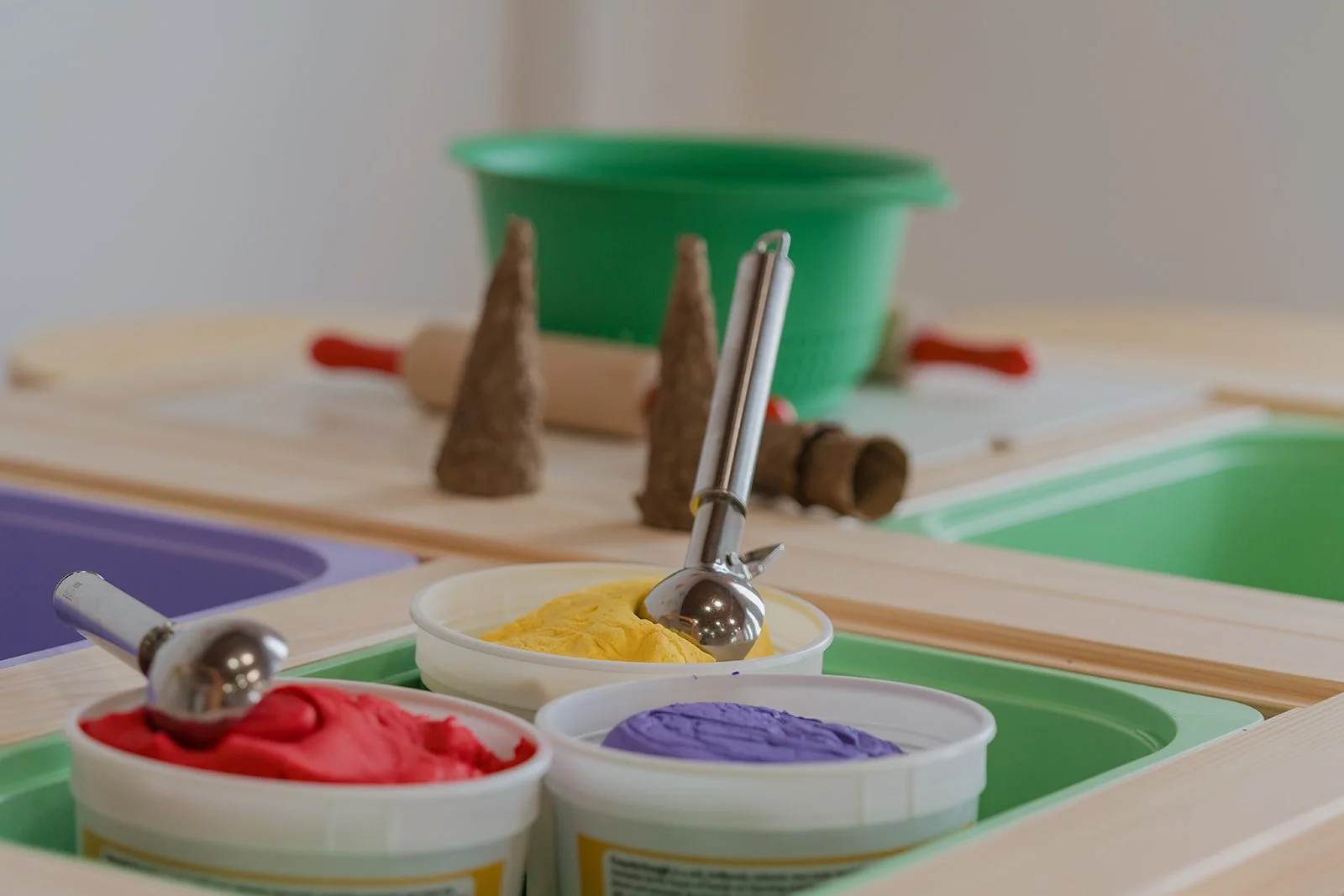 Colorful ice cream tubs with scoops, a green container in the background, and some miniature tree and log toys on a wooden surface.